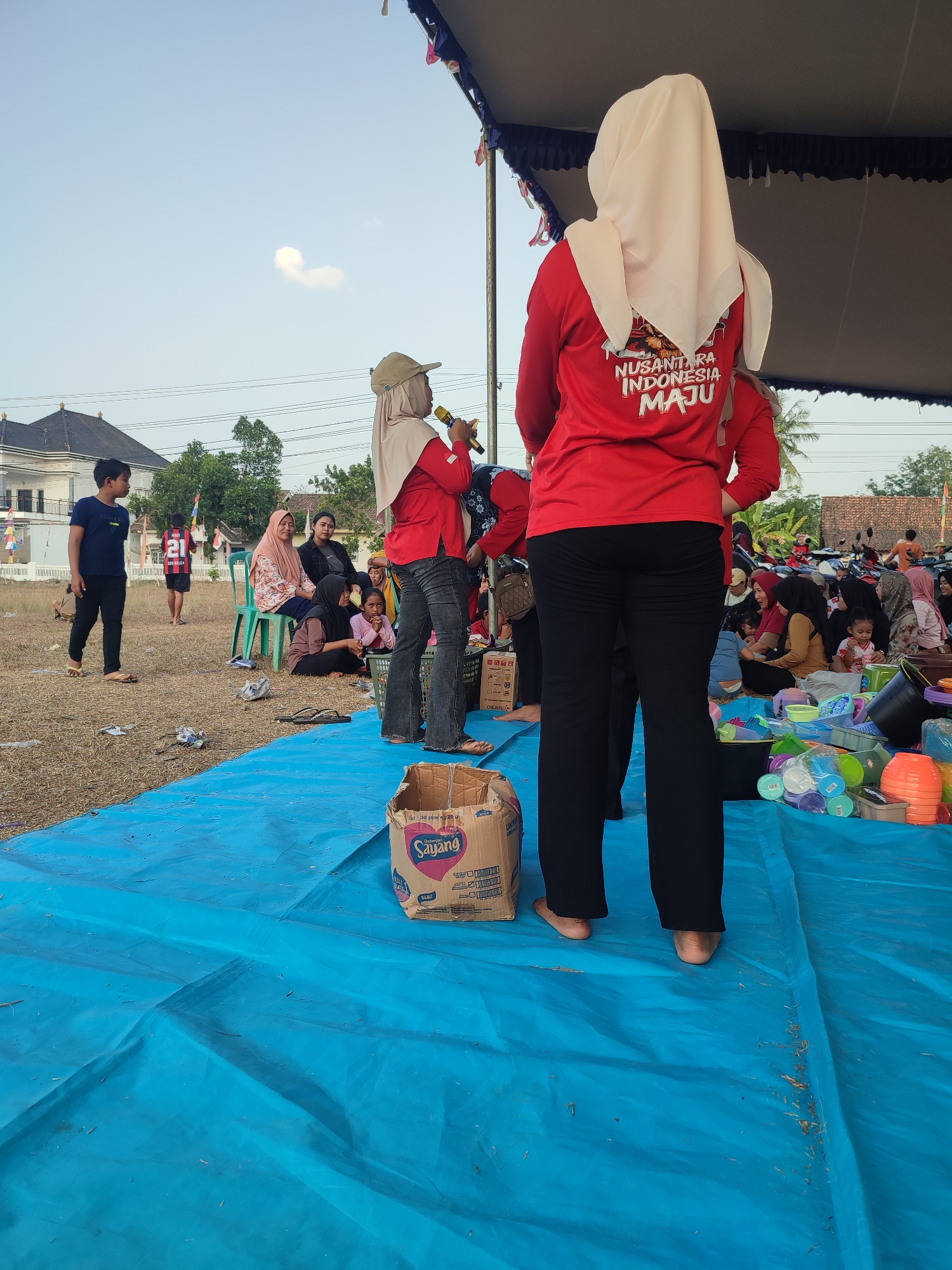 An outdoor event is taking place in Nusa Jaya, Indonesia, under a large tent. The ground is covered with a blue tarp, with dry grass and debris visible in some areas. A group of people, mostly women and children, are gathered on the ground and sitting on chairs. Some are wearing hijabs, and the attire suggests a casual, community-oriented gathering.
In the foreground, a woman wearing a red shirt with text "NUSANTARA INDONESIA MAJU" and black pants, with a light-colored hijab, holds a microphone and appears to be speaking. Another person in a similar red shirt stands beside her. To the left, a young boy in a dark blue t-shirt and black pants walks across the blue tarp.
Further back, several women are seated on the ground and on green plastic chairs. Among them, one woman in a pink floral patterned top sits on a chair, and others are seated on the ground, some holding or interacting with children. In the background, a building with a dark roof and white walls is visible, along with trees and other structures. A row of motorcycles is parked in the distance, indicating a populated area.
Various objects are spread around the scene, including plastic containers and buckets of different colors, suggesting that the event might involve distribution of goods or some form of activity. A cardboard box with the brand "Sayang" is also prominently displayed on the blue tarp in the foreground. The overall atmosphere appears to be active and social, possibly a local festival or community outreach program.