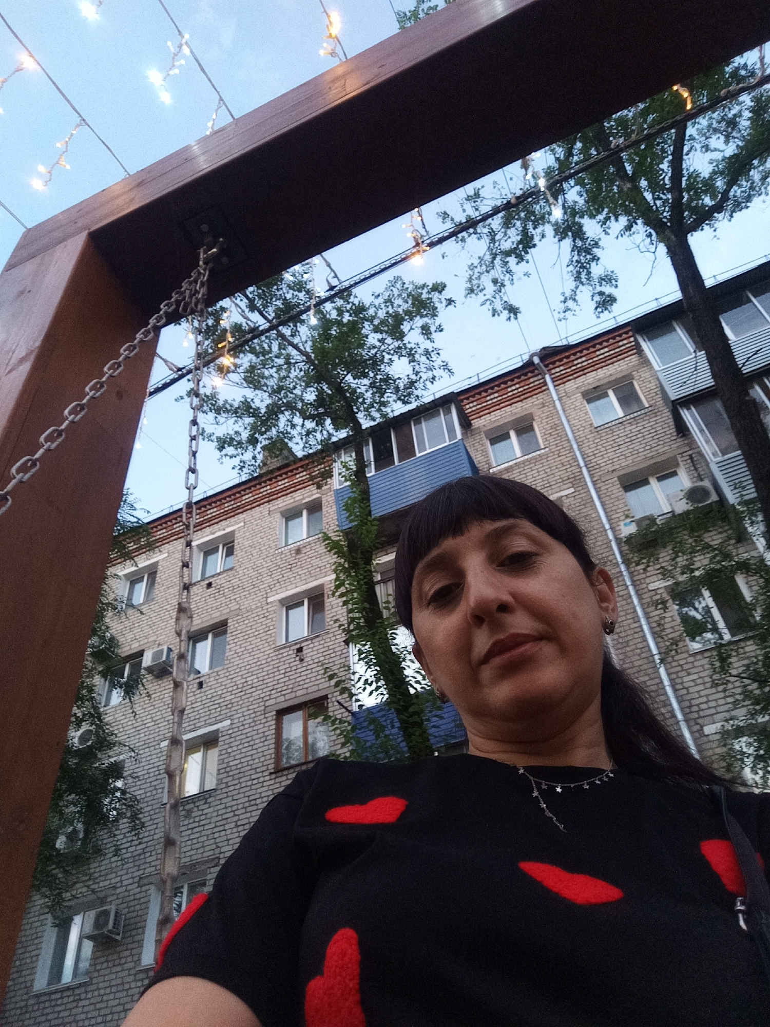 A woman is taking a selfie outdoors in front of a brick apartment building. She is wearing a black shirt with red accents and a silver necklace. A wooden structure with chains and string lights is visible in the foreground. Trees are present, and the sky is a light blue. The location is the city of Belogorsk, Russia.