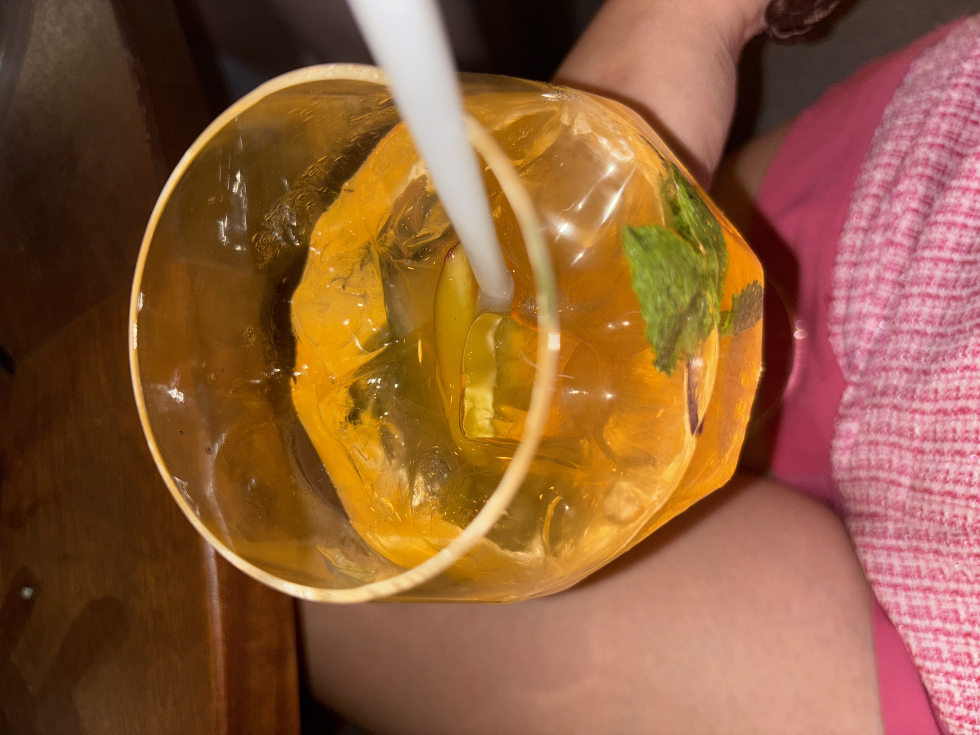 A close-up, top-down shot focuses on a large, clear glass filled with an amber-colored beverage and ice cubes. A white straw is inserted into the drink, positioned to the right of center. A few sprigs of fresh mint and a slice of citrus are visible within the glass. The drink is condensation-free and illuminated from above, creating subtle reflections on the glass surface. The background shows a portion of a dark wooden table and the lower half of a person's body, clad in bright pink clothing and a pink and white textured garment. The overall setting appears to be indoors, possibly at a restaurant or bar, given the ambient lighting and table surface.