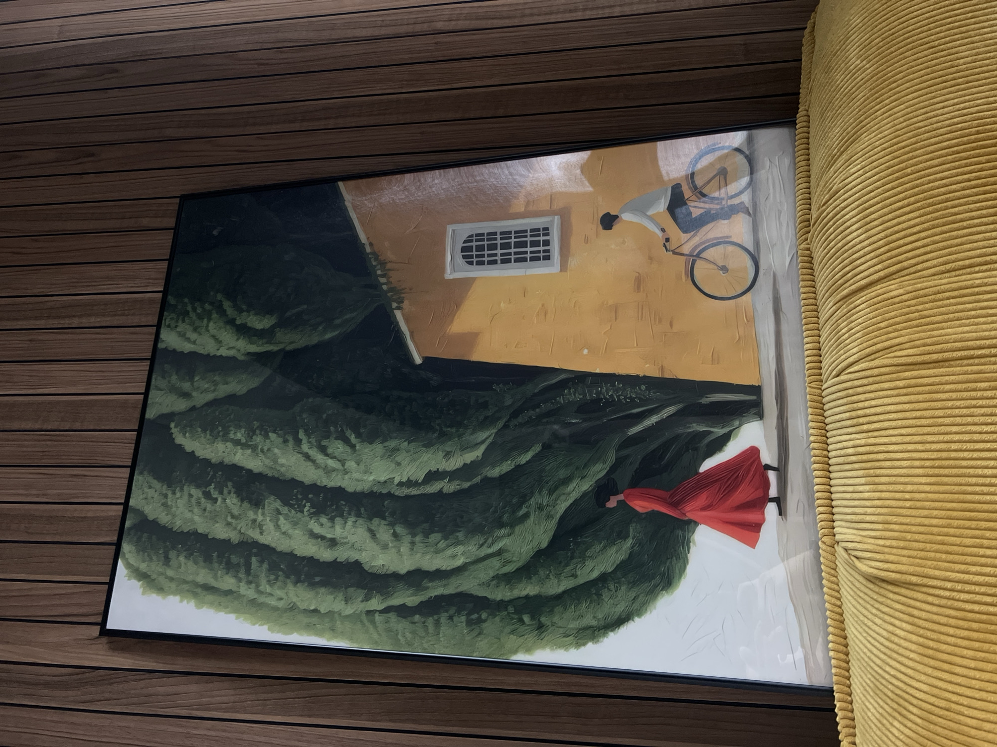 This is a photograph of a painting hanging on a wall with vertical wooden paneling. The painting depicts a scene with a large, dark green tree on the left, partially obscuring a yellow building on the right. A woman in a flowing red dress is walking on a path in front of the tree, and a person on a bicycle is riding on a path in front of the building. The building has a white-paned window. The bottom of the image shows a ribbed, yellow fabric surface. There is no indication that this scene is located in the city of Penza, Russia.