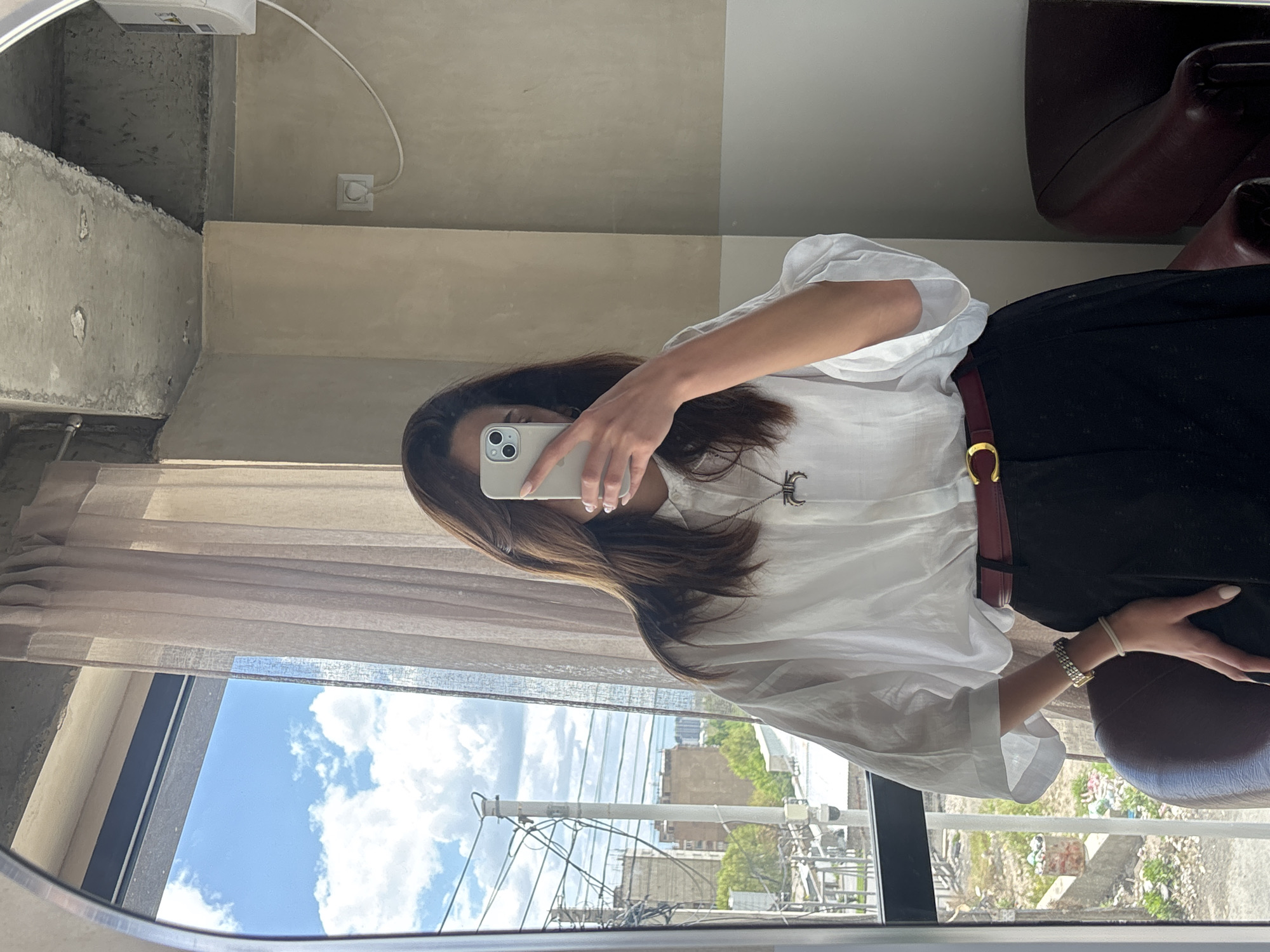 The image is a selfie taken by a woman in front of a mirror. She is wearing a white blouse, a dark skirt, and a burgundy belt with a gold buckle. The woman is holding a white smartphone with two cameras in her left hand, partially obscuring her face. The reflection shows a window with a view of a city street, including utility poles, buildings, trees, and a blue sky with scattered clouds. The location appears to be Yerevan, Armenia, based on the visible architectural style and the appearance of utility infrastructure. Inside, a portion of a dark-colored chair is visible, along with a gray ceiling. A sheer curtain partially covers the window.