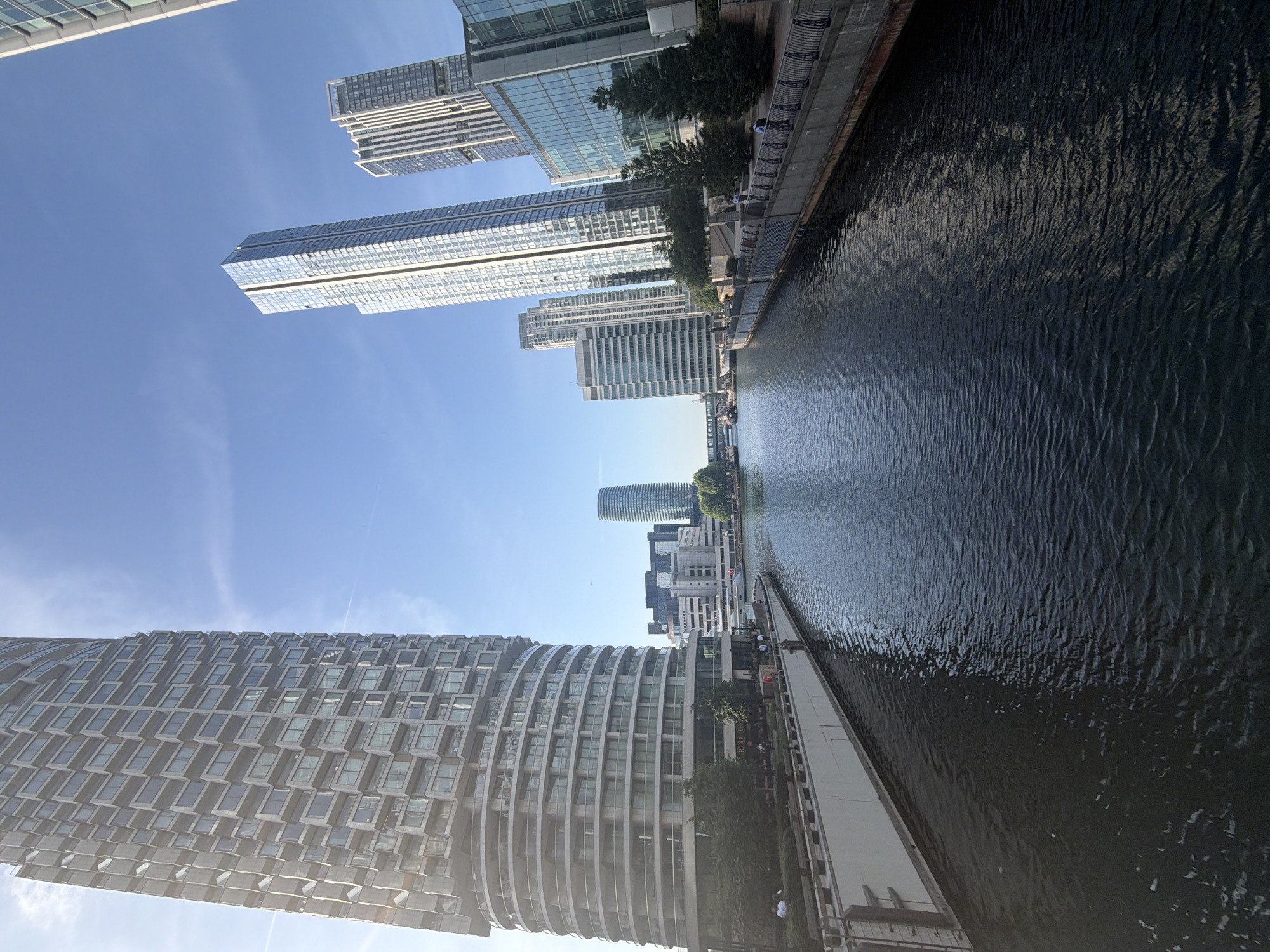 The image shows a view of the Canary Wharf area in London, United Kingdom. A body of water reflects the sky and some of the buildings. The buildings are tall and modern, with glass facades. There are a few people visible along the waterfront. The weather appears sunny.