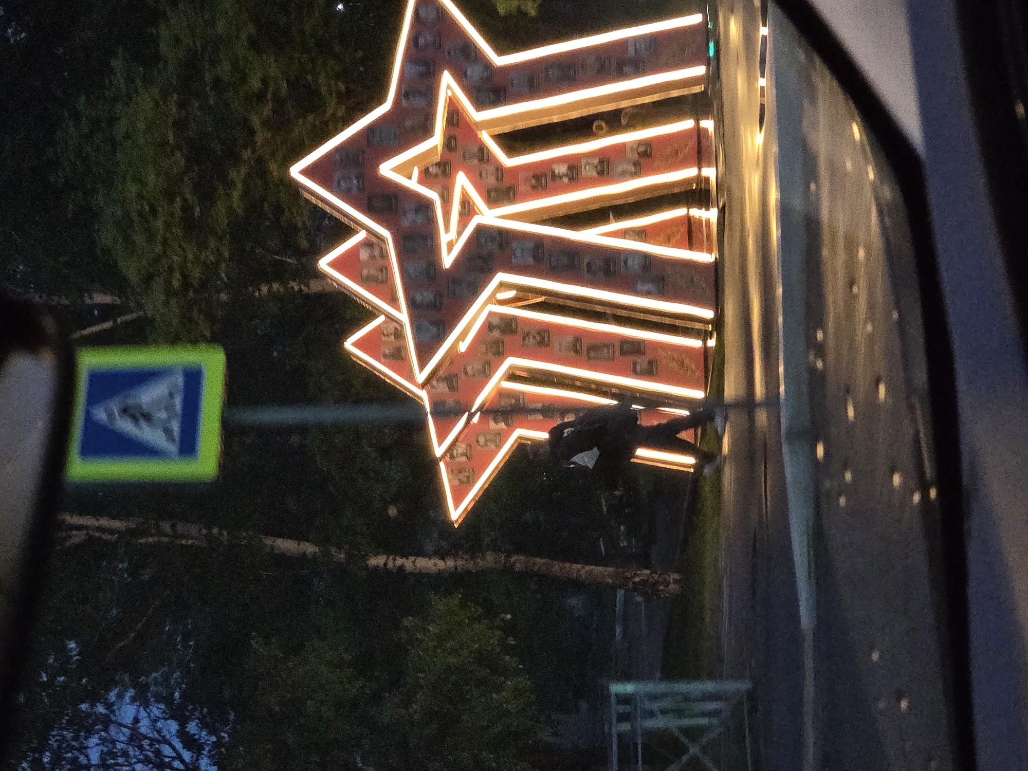 The image depicts a nighttime scene in Birobidzhan, Russia. The focus is on a large, illuminated three-dimensional structure comprised of three red stars with yellow edging. The stars are decorated with numerous small photos. To the left of the stars is a pedestrian crossing sign, while trees are visible in the background. In the middle of the image, a person is visible near the star structure. The image appears to be taken through a window, possibly of a car, as indicated by the reflections on the right.
