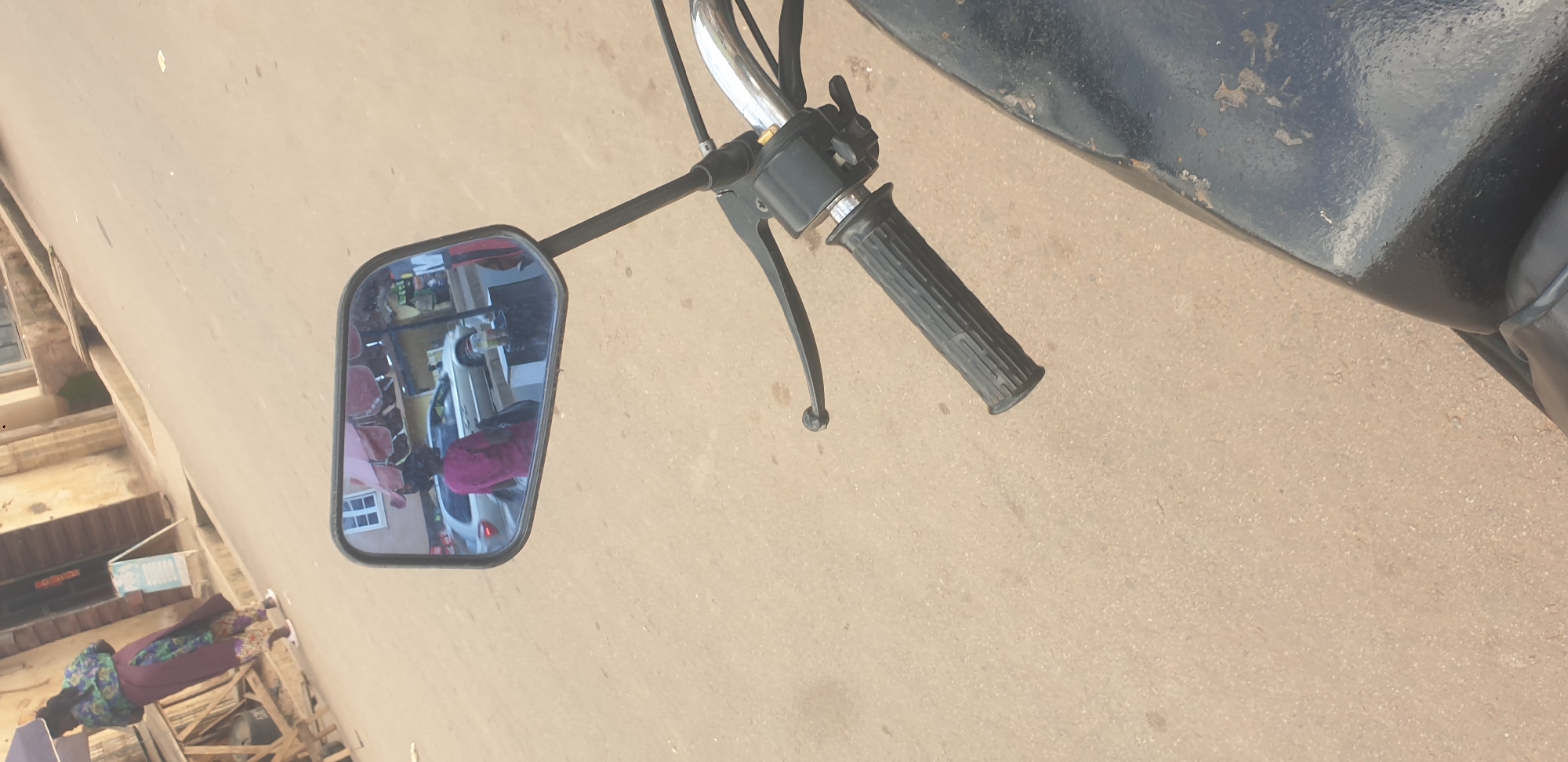 The image captures a street scene in Barakin Lerawi, Nigeria, as seen from the perspective of a motorcycle. The focus is on the motorcycle's handlebar and a rearview mirror. The mirror reflects a street with buildings, people, and vehicles. A woman in a pink shirt and dark skirt is walking away from the camera, with a silver car behind her and a market stall to the right. The street is paved with dirt or gravel. The motorcycle's black handlebar is visible in the foreground.
