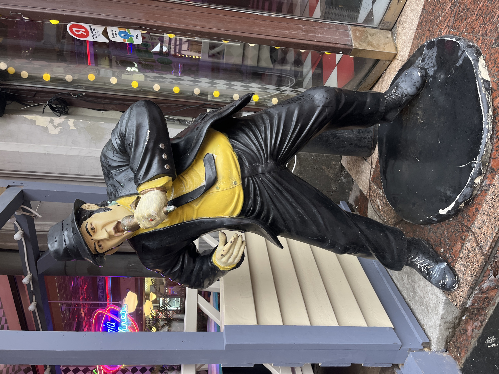 A life-sized statue of a man stands outside a building. He wears a black suit with a yellow shirt and a black fedora, holding a cigar. The statue appears to be made of painted resin or fiberglass. He stands on a black platform. The background includes a storefront with a window displaying neon signs and checkered patterns. A sign in the window has Cyrillic text, indicating a location in Moscow, Russia. The statue is placed on a sidewalk near the entrance of what appears to be a restaurant or business.