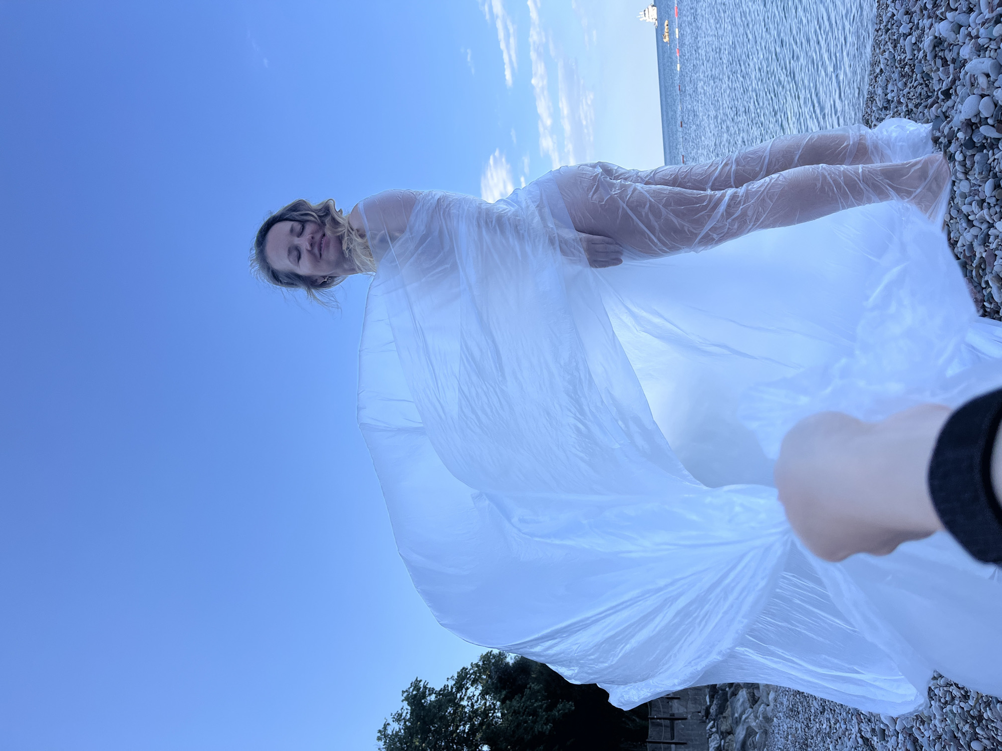 The image, taken in Boreti, Montenegro, presents a woman on a pebble beach, partially covered by what appears to be a large, transparent plastic sheet or wrap. The woman, seemingly lying down, faces the camera with her eyes closed. Her blonde hair is loose. She is on a pebble beach that is visible in the background, along with the water and horizon. The sky is a clear blue. A hand wearing a black sleeve is visible, holding the plastic wrap. In the bottom part of the image, we can see trees and a barrier. There is a ship on the sea's horizon. The focus is on the woman, with the plastic wrap being a significant element in the frame.