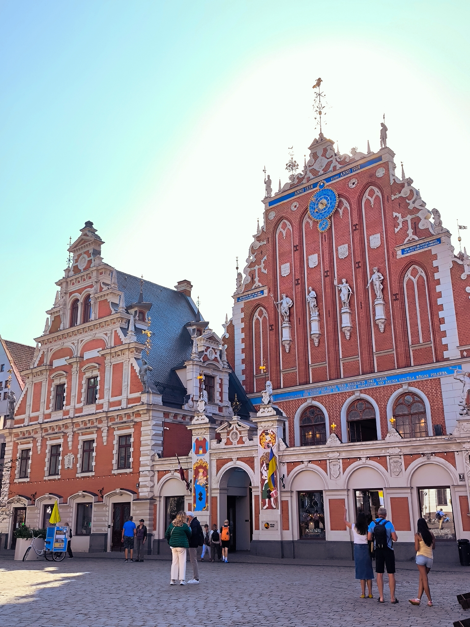 The image shows a view of the House of the Blackheads in Riga, Latvia, a historic building located in the Old Town. The facade is adorned with intricate architectural details, including statues and decorative elements. A large blue clock is prominently displayed on the front of the building. In the foreground, several people are walking on a cobblestone square. The sky is clear and bright, indicating a sunny day. The overall scene depicts a bustling urban environment with historical significance.