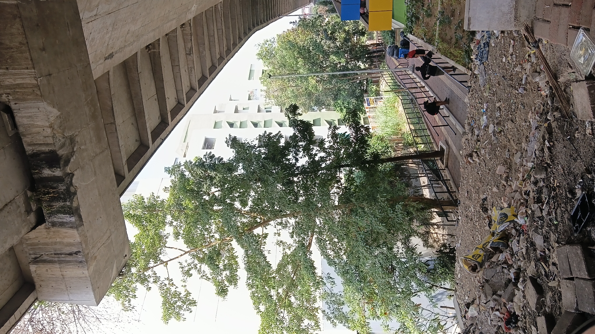 The image captures a scene under a concrete overpass in Bandung City, Indonesia. In the foreground, a pile of rubble and construction debris, including broken bricks and rebar, is visible. A yellow, broken object resembling a scooter or a shareable bike lies among the debris. A paved pathway with a black metal railing winds upwards in the mid-ground. Three individuals are present on this pathway: one person is walking uphill, another is stopped and appears to be looking down, and a third person is walking downhill carrying a red bag. Beyond the pathway, a modern white building with green triangular accents on its facade is situated, surrounded by lush green trees. The overpass structure is a prominent feature, with its rough concrete underside and supporting beams. The overall impression is one of urban infrastructure juxtaposed with a somewhat neglected and cluttered foreground, with a hint of everyday life in the mid-ground.