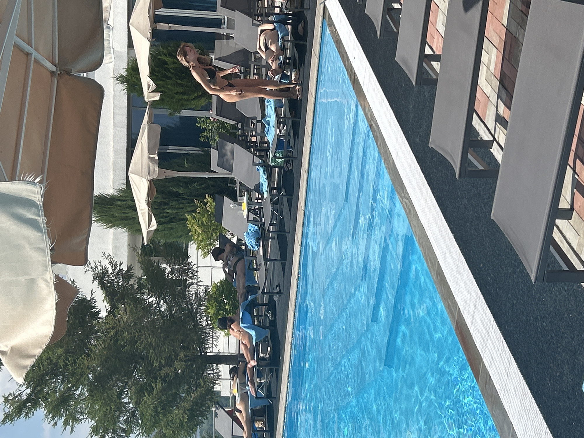 A group of people are relaxing by a bright blue swimming pool on a sunny day. The camera is rotated 90 degrees counter-clockwise, so the scene appears sideways. In the foreground, a woman in a black bikini stands on a lounge chair, leaning back and looking up, with her arms extended behind her. Several other individuals are seated on lounge chairs on the deck of the pool, some with towels. The pool is covered with a blue tarp.
Behind the pool, there are lounge chairs, trees, and part of a building with large windows. Umbrellas with light-colored canopies are also visible, providing shade. The ground around the pool is paved with dark material, and the pool's edge is lined with white and beige tiles. The overall atmosphere appears relaxed and leisurely, suggesting a vacation or a day off. The time of day is clearly daytime, indicated by the strong sunlight and shadows. The location is identified as Novokuybyshevsk, Russia, but there are no specific visual cues to confirm this within the image itself. No text is visible in the media file.