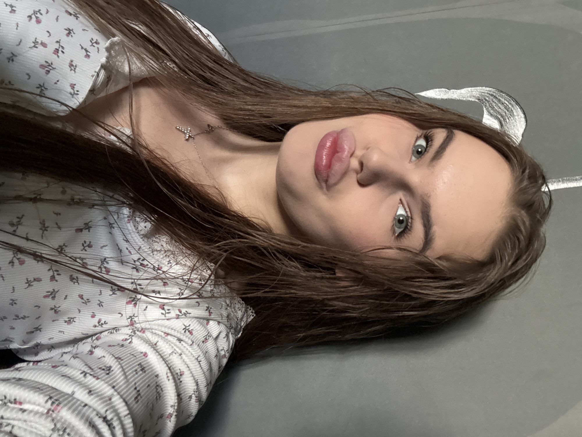A young woman with long, wet, brown hair and blue eyes is looking at the camera, pursing her lips. She is wearing a white top with a floral pattern and a cross necklace. The background is a gray wall with a faint white design. The image appears to be a selfie taken indoors. There is no information in the image that indicates the location in the city of Dnipro, Ukraine.