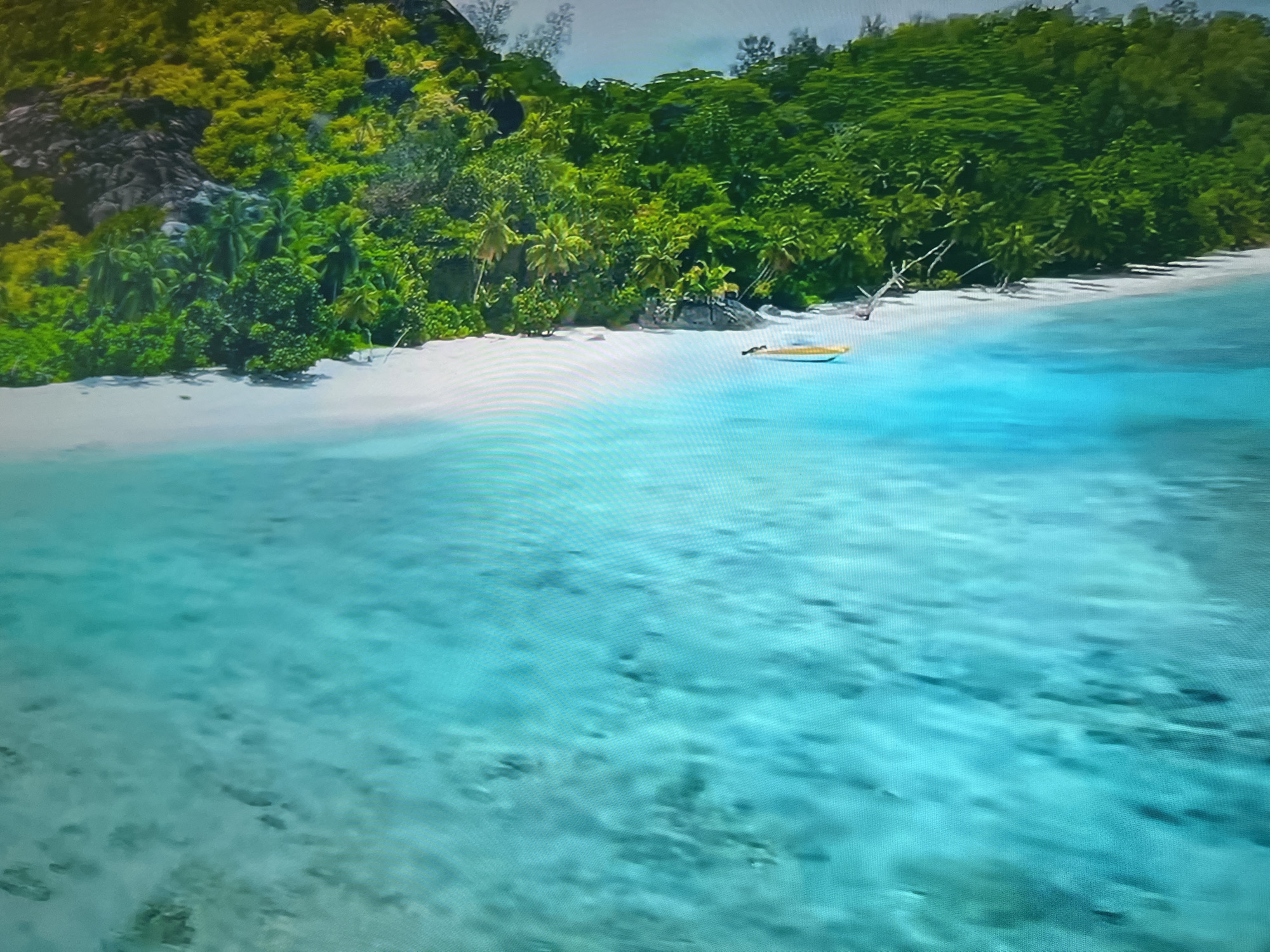 The image depicts a tropical beach scene. Lush green vegetation covers the land, transitioning to a white sandy beach. Turquoise water extends from the beach into the foreground. A yellow kayak rests on the sand near the shoreline. No people are visible. The location is not within Basingstoke and Deane, United Kingdom.