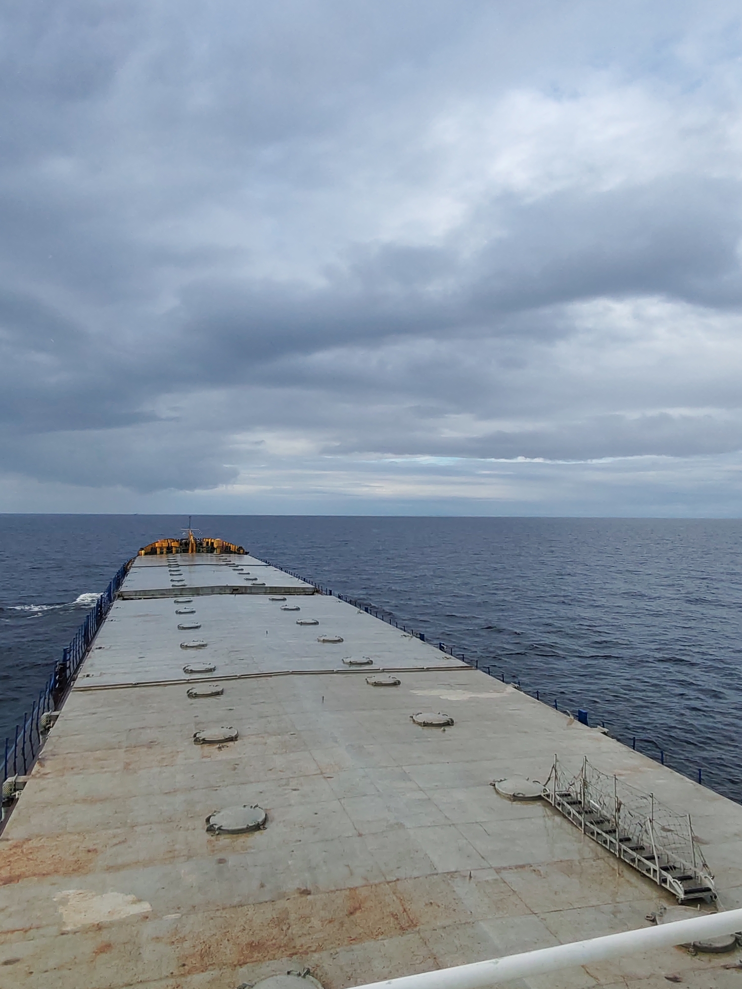 The image shows the deck of a large ship at sea. The deck is gray and metallic, with numerous round hatches. A set of stairs or a gangway is visible on the right side of the deck. The ocean is a dark blue-gray, and the sky is overcast with heavy, gray clouds. The location is unknown, Russia is not known.