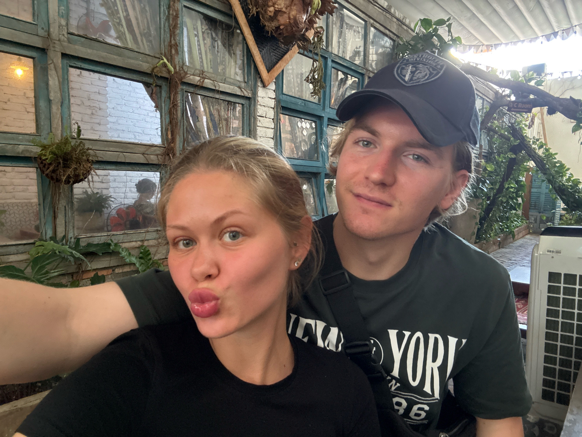 A close-up selfie shows a young Caucasian couple posing in front of a windowed wall. The woman, in the foreground, puckers her lips for the camera. She has blonde hair pulled back and is wearing a black t-shirt. The man, behind her and to her right, smiles and wears a dark baseball cap and a dark green t-shirt with white text that reads "YORK 96". The background is a rustic outdoor setting with a windowed wall featuring teal-colored frames and reflections of a brick building. Hanging plants and vines are visible, along with a decorative banner strung across the ceiling. To the right of the couple, a white air conditioning unit is positioned. The lighting suggests it is daytime. The overall ambiance is casual and relaxed.