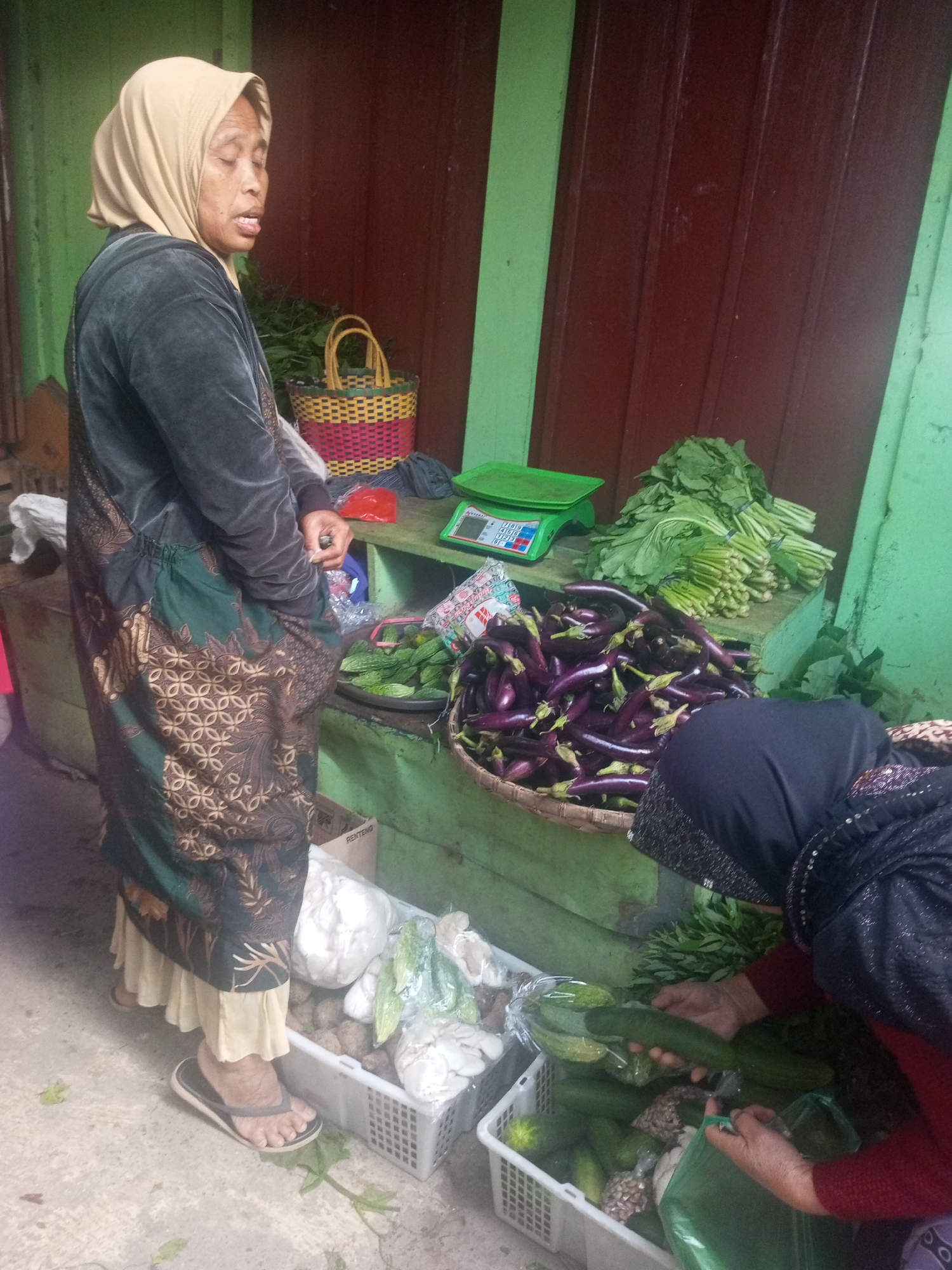 The image captures a scene at a market stall in Kalibeber, Indonesia. In the foreground, a woman wearing a light brown hijab and a dark velvet-like robe with a patterned fabric over a pale yellow skirt stands facing left. She is wearing flip-flops. To her right, another person, partially visible and wearing a dark hijab and a maroon top, is reaching into a white plastic basket filled with cucumbers. This basket is placed next to another white plastic basket containing what appear to be leafy vegetables and possibly onions or garlic. In the middle ground, a large woven basket is overflowing with purple eggplants. Behind the eggplants, a pile of green leafy vegetables is arranged on a green surface. To the left of the eggplants, a brightly colored woven basket with red and yellow sections sits on a shelf. A green digital scale is visible on the shelf, next to a red object, possibly a bag. The stall appears to be constructed with green and dark red wooden planks. The ground beneath the woman's feet is concrete, with some scattered green leaves.