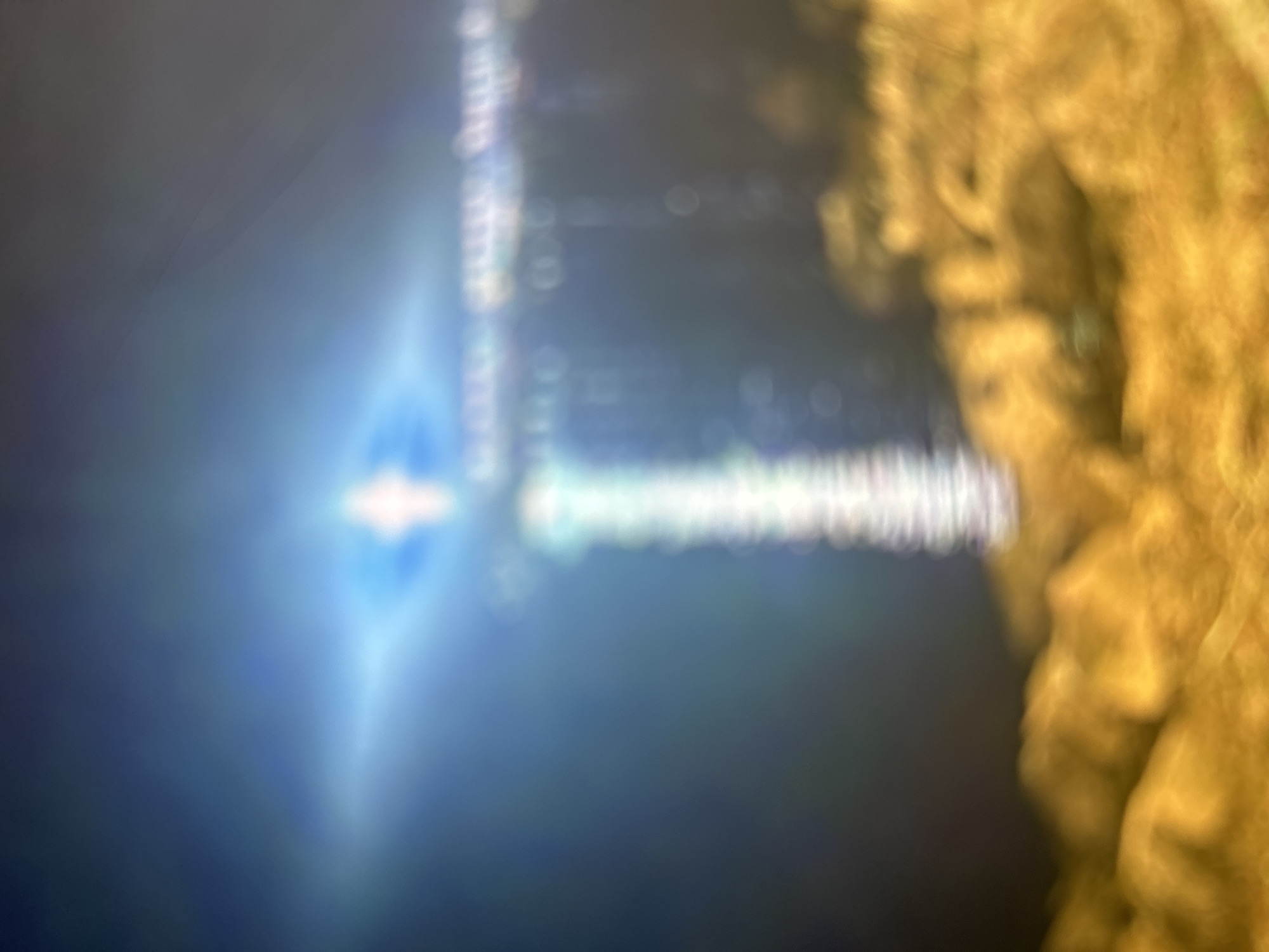 The image shows a blurry scene. On the left, there is a bright blue light with a starburst effect. Moving towards the right, there's a streak of lighter colored bokeh shapes. The right side of the image is dominated by a blurry, yellow-brown mass that appears to be organic in nature. The location is listed as Unknown, Unknown. No people, clear objects, actions, or interactions are discernible.