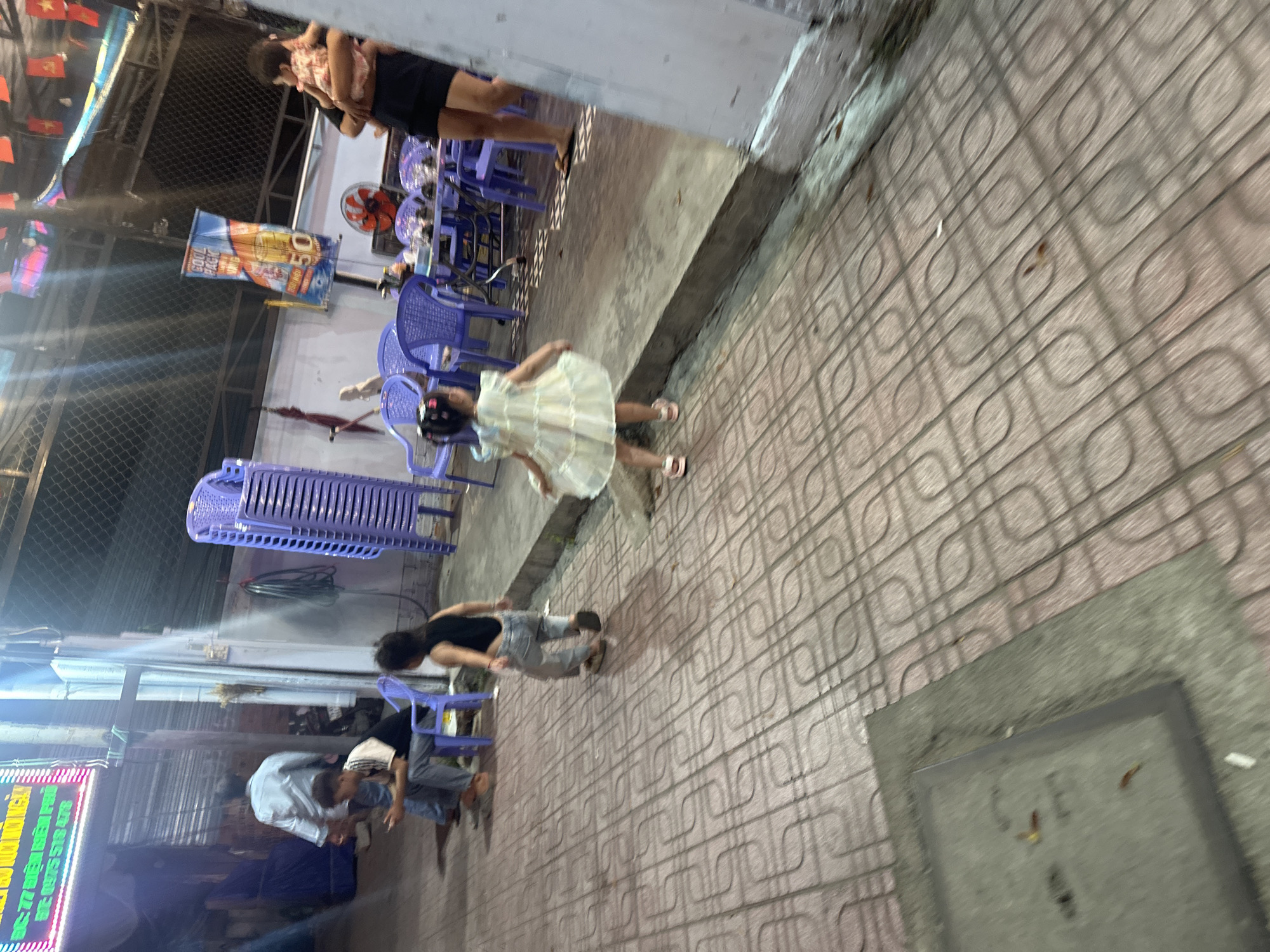 The scene is a nighttime streetscape in Bac Nha Trang Ward, Vietnam. A child in a white dress with a black helmet is standing on the sidewalk. Several adults stand nearby or sit on purple plastic chairs. Stacks of the same chairs are present near the wall, and some people are gathered near a lit-up sign. A banner is hung in the background. The sidewalk is tiled with a repeating loop pattern.