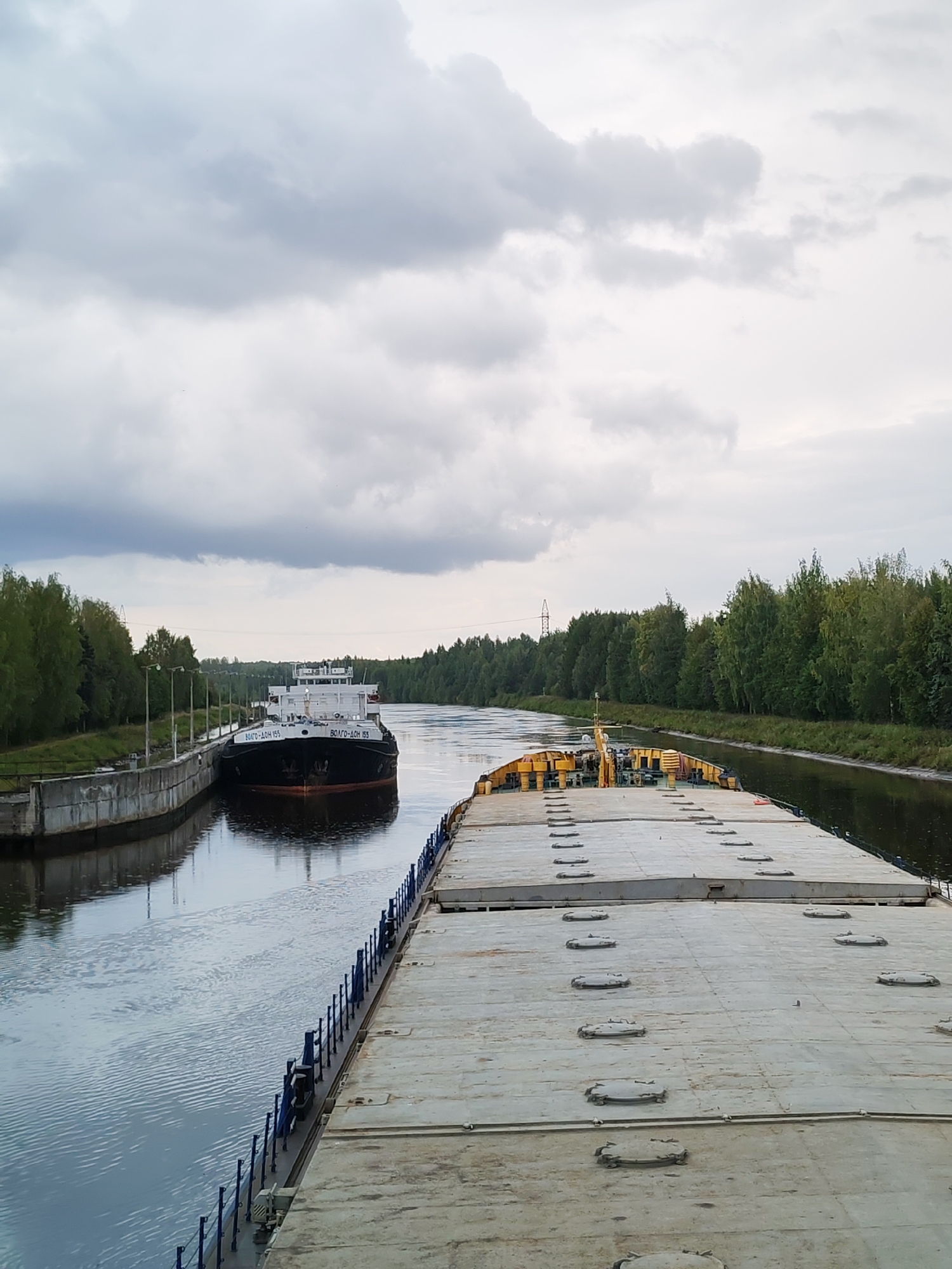 An overcast day is depicted in Vytegra, Russia, as two barges navigate a canal. The foreground is dominated by the deck of a large, flat barge, its surface marked by numerous circular hatches. In the midground, a second barge, a dark-hulled vessel with white superstructures, is docked alongside a concrete structure. The canal is flanked by lush green trees on both sides, with a power line visible in the distance. The water reflects the cloudy sky and the surrounding vegetation.