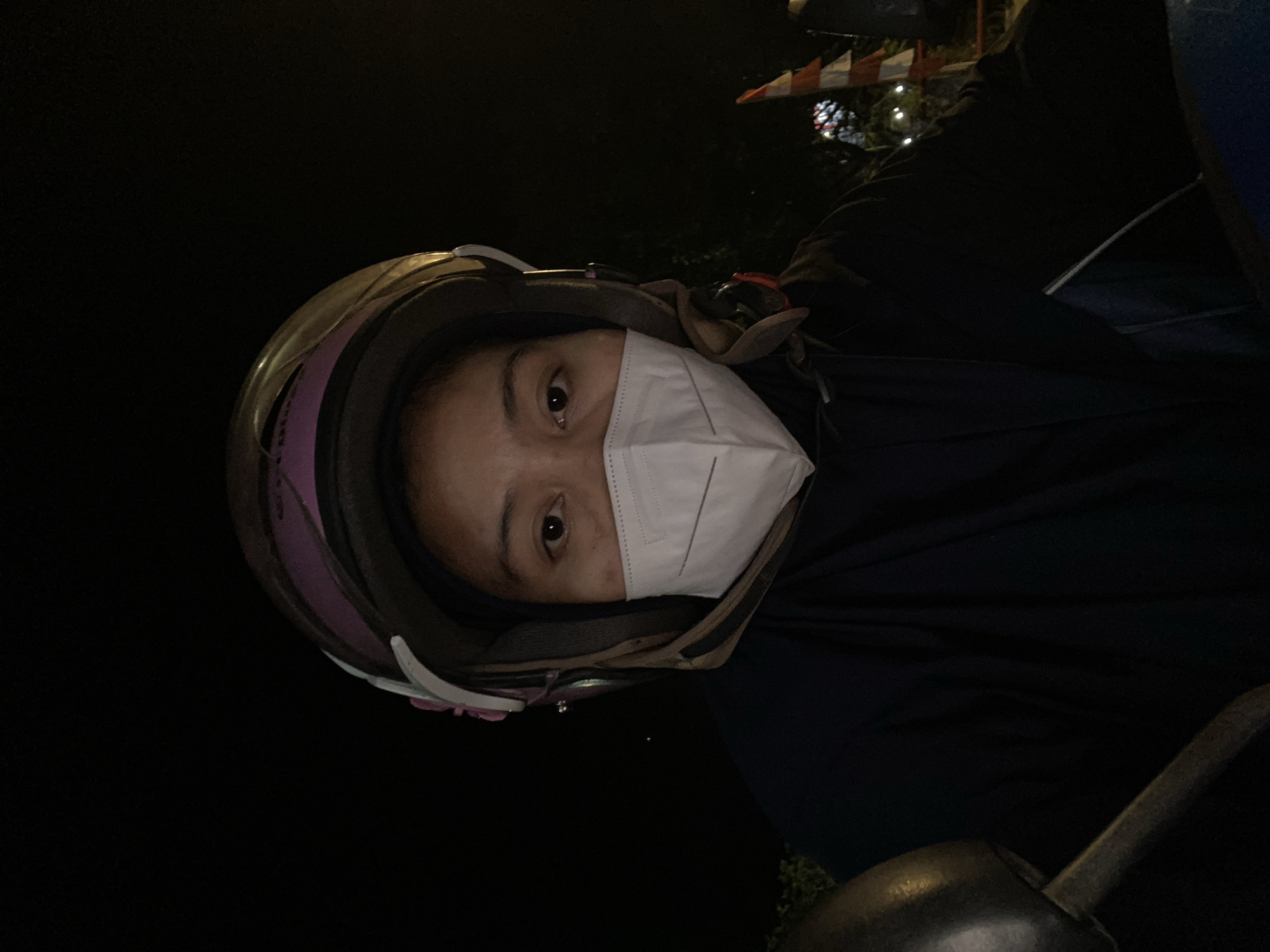 A first-person perspective selfie taken at night shows a person wearing a motorcycle helmet and a white surgical mask. The person's eyes are visible above the mask. The background is dark, with faint lights and trees visible in the distance, suggesting an outdoor urban setting. The person appears to be on a motorcycle, with a portion of the handlebars visible in the lower left corner of the frame. The scene is likely in Gambut Barat, Indonesia, given the context.