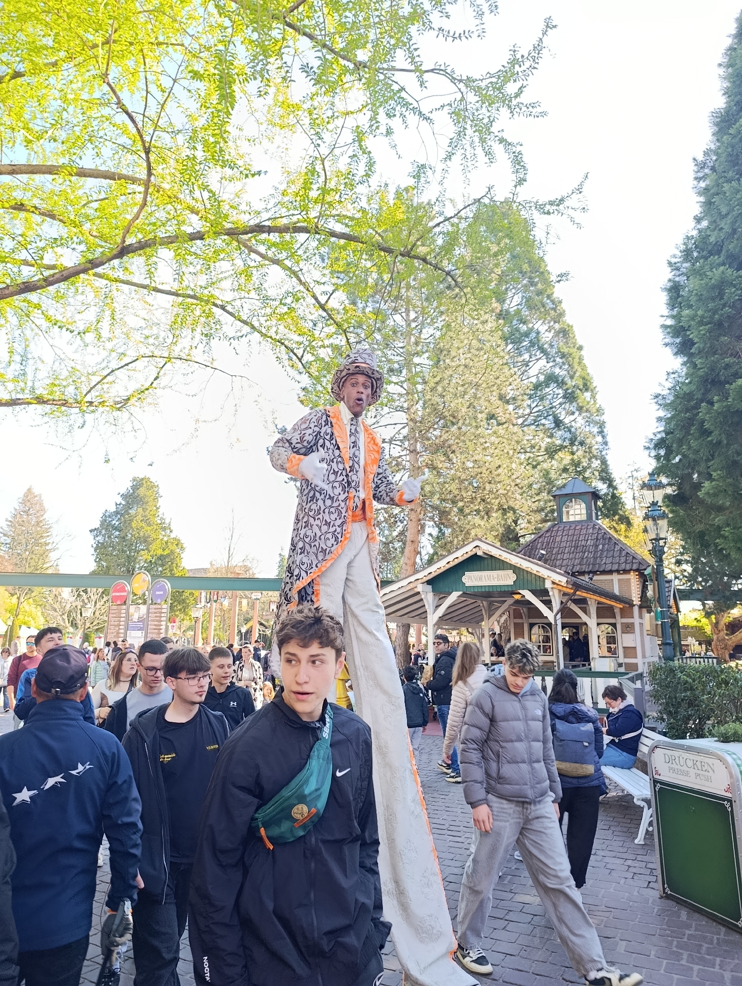 The image depicts a street scene in Rust, Germany, likely within or near the Europa-Park amusement park, due to the presence of the "Panorama-Bahn" station in the background. A person on stilts, dressed in a patterned coat, white pants, and a hat, is the central focus, performing or interacting with the crowd. The crowd consists of a diverse group of people, including teenagers and adults, some of whom are looking at the performer. The foreground features individuals walking towards the viewer. The environment includes trees, buildings resembling park structures, streetlights, benches, and signage (one indicating "Drücken Presse Push"). The lighting is bright, suggesting daylight, with tree branches and leaves visible overhead.
