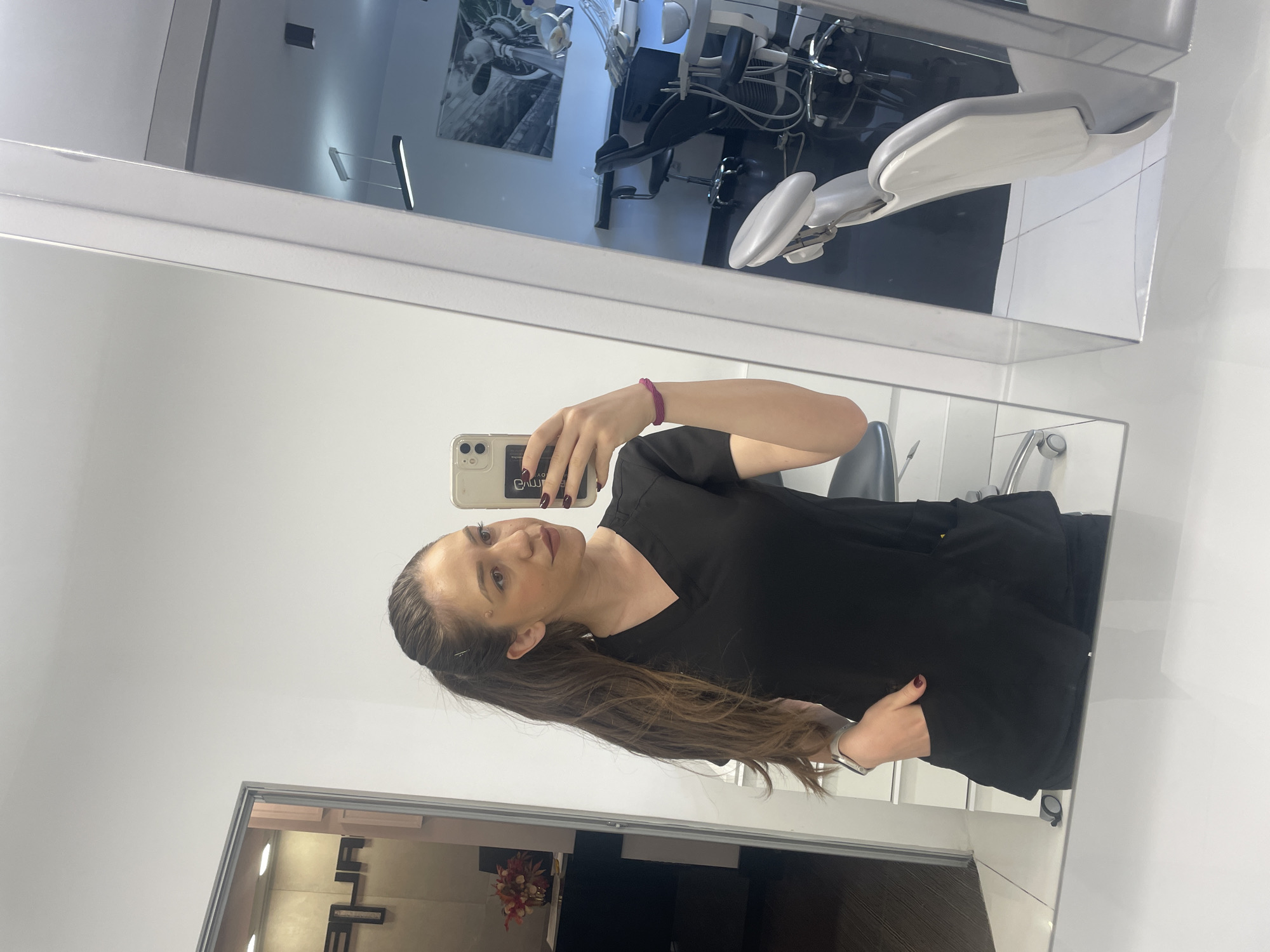 The image shows a woman taking a selfie in a mirror. She is wearing a black top and has long brown hair. She is holding a cell phone in her left hand. The background of the image shows a dental office. A dental chair is visible in the mirror's reflection. The environment appears clean and modern. There is no explicit indication of the city of Sofia, Bulgaria in the scene.