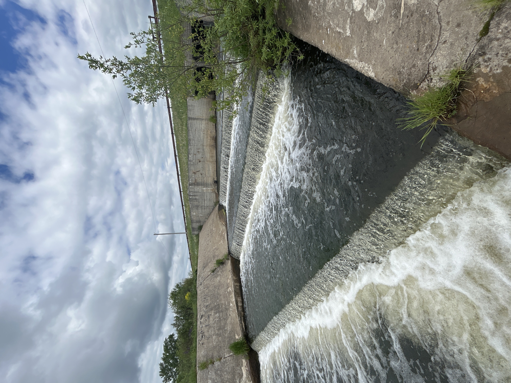 The image depicts a tiered concrete dam structure with water cascading down each level. Vegetation, including trees and grass, grows along the dam's edge and around the concrete. The sky is overcast with a mix of light and dark clouds. The location is identified as Большая Уса, Russia. There are no people visible in the scene.