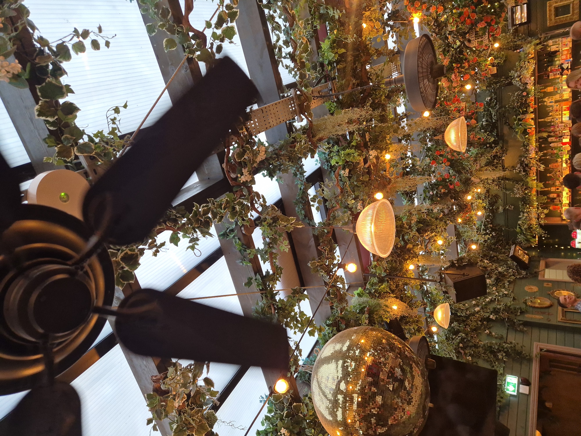 The image shows a ceiling decorated with lush greenery and various light fixtures, likely inside a bar or restaurant. The ceiling structure consists of wooden beams and translucent panels. Two ceiling fans are visible, one prominently in the foreground. Strings of warm-toned lights and decorative lamps are intertwined with the foliage. A large disco ball hangs towards the lower part of the image. In the background, a wall is visible, also covered in greenery, with bottles and decorative items arranged on shelves. This scene is likely located in The Botanist in Cardiff, United Kingdom. People are indistinctly visible near the wall.