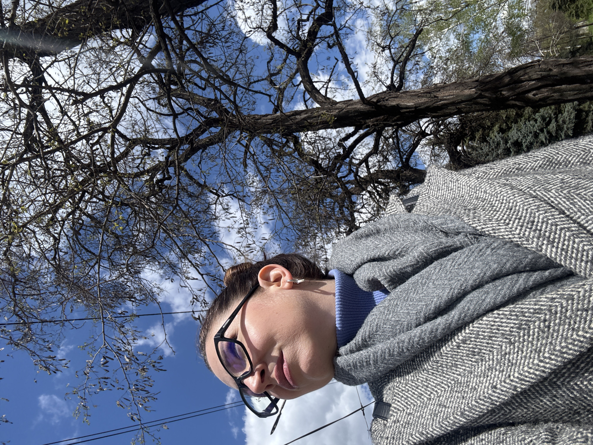 A person is looking upwards at a tree against a partly cloudy sky. The person is wearing glasses and a gray coat and a blue turtleneck sweater. The tree is largely bare of leaves. The location is unknown, there is not enough information to determine it is in Kryvyi Rih, Ukraine.