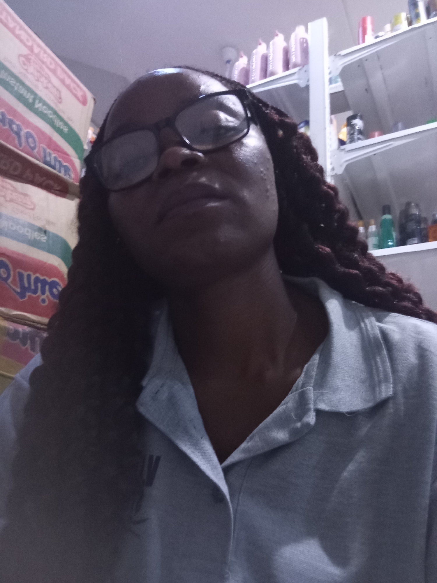 This is a medium shot of a young Black woman wearing glasses and a light blue collared shirt. She is looking upwards and to her left. The woman has dark skin and her hair is styled in long, dark braids. She appears to be in an indoor setting, possibly a store or a home, with shelves filled with various products in the background. The shelves on the left are stacked with what appear to be boxes of food items, possibly noodles, with text visible in a foreign language. The shelves on the right are stocked with bottles and containers, likely personal care or cleaning products. The lighting is dim, suggesting it may be evening or nighttime. The overall mood is neutral. There is text visible on the boxes of food on the left, some of which appears to be in a script that is difficult to read due to the resolution and angle.