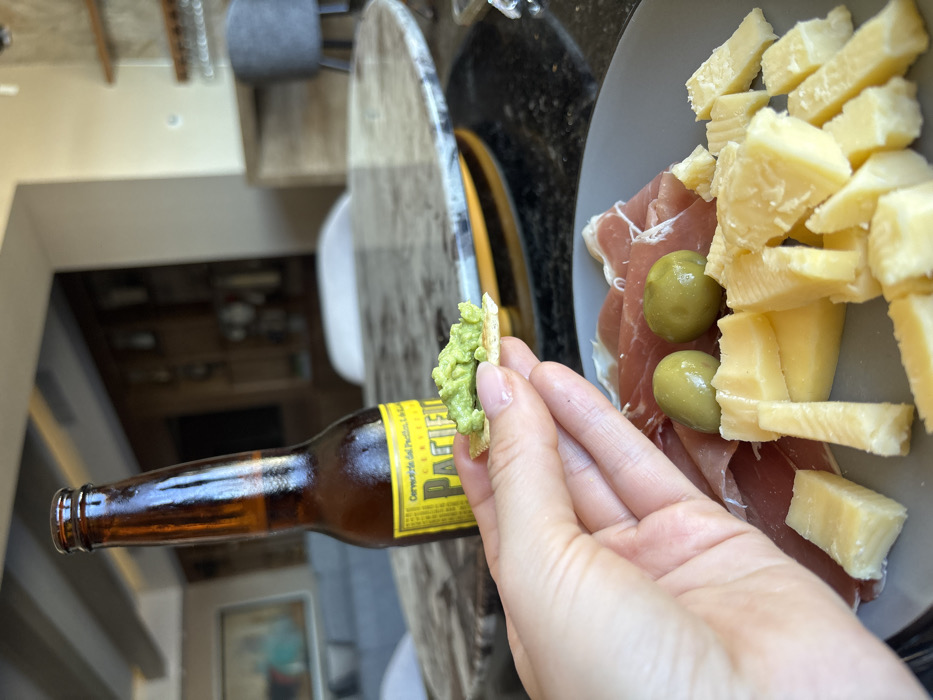A hand holds a cracker topped with green dip next to a bottle of Pacifico beer. A gray plate on a counter features thin slices of prosciutto, green olives, and chunks of Parmesan cheese. The counter is black with a patterned edge. A glimpse of the interior of a house is visible in the background.