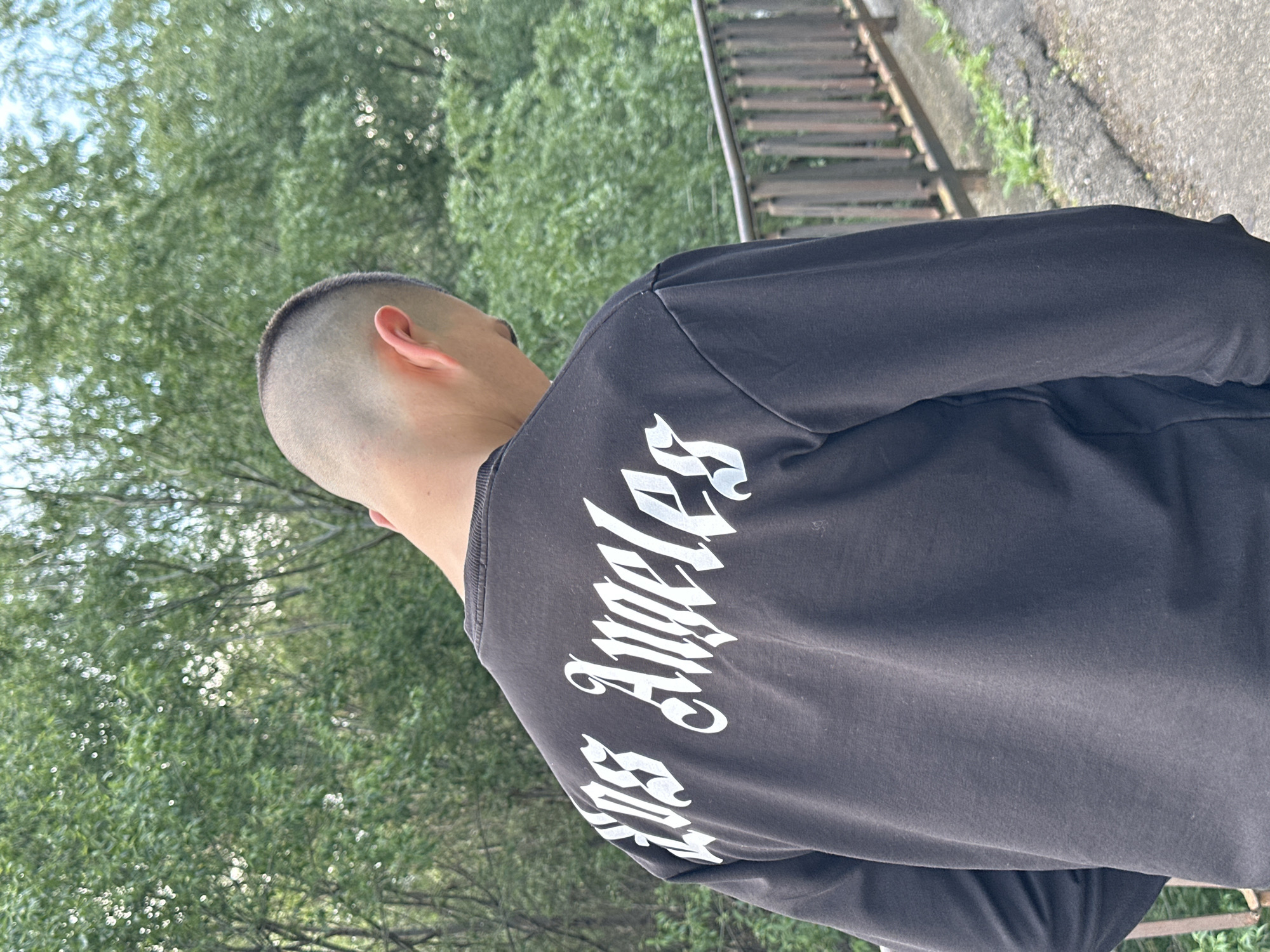 A person with a shaved head and a black long-sleeved shirt is standing near a set of railroad tracks. The shirt has the words "Los Angeles" printed on the back in white gothic-style font. The person's back is to the camera, and they appear to be facing a green, leafy area. The scene seems to be outdoors. The location is Lovech, Bulgaria.