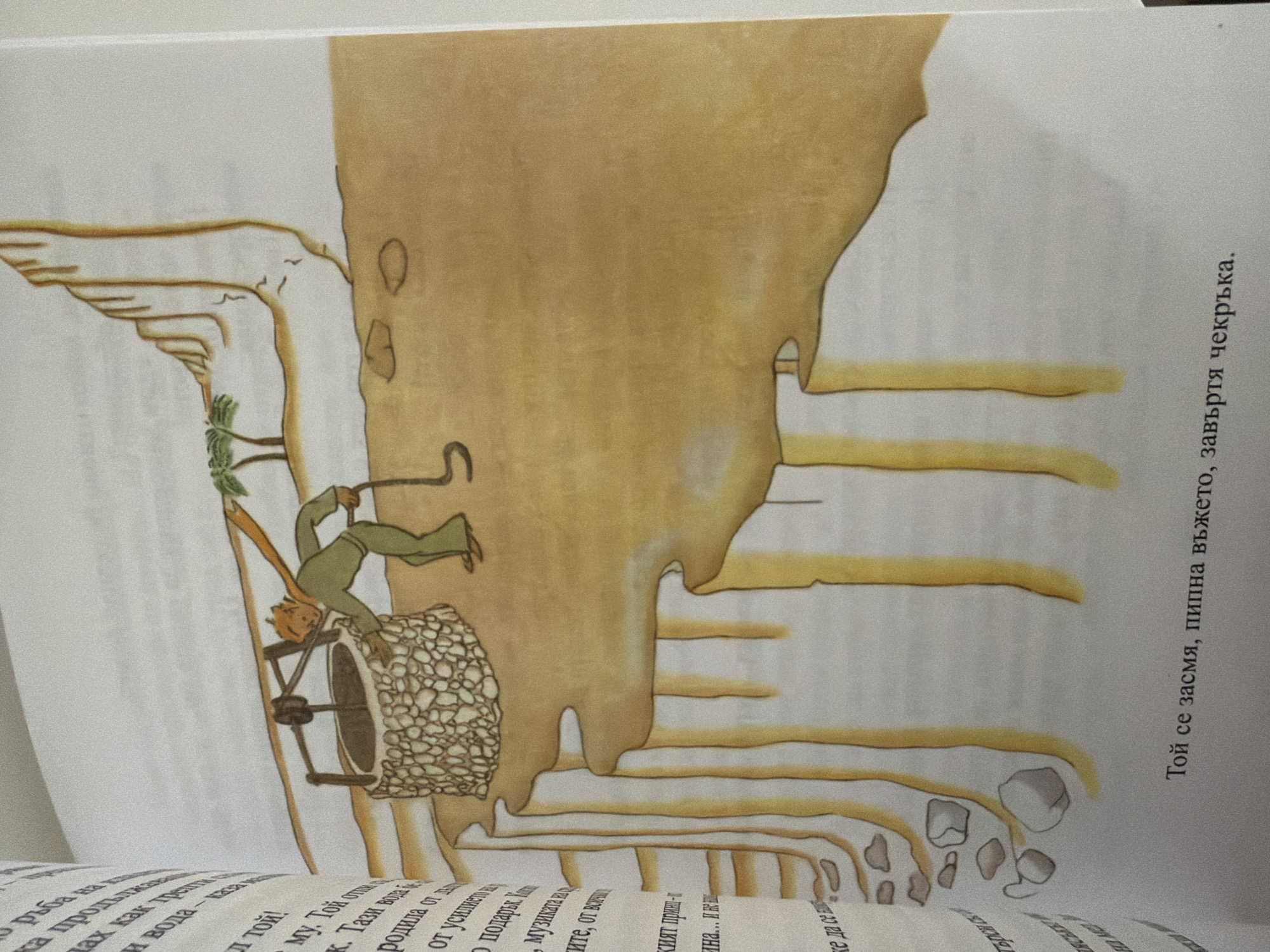 The image depicts an illustration from a book, showing a character dressed in green, leaning over a stone well. The character is holding a rope attached to a crank mechanism. The well is situated on a sandy landscape with a rocky cliff in the background and a small palm tree. Below the well, the sand appears to be eroded, forming deep channels. The text at the bottom of the image is in Bulgarian and reads, "He sighed, and turned the crank." There is no indication or visual element within the image that suggests a location in Pazardzhik, Bulgaria.