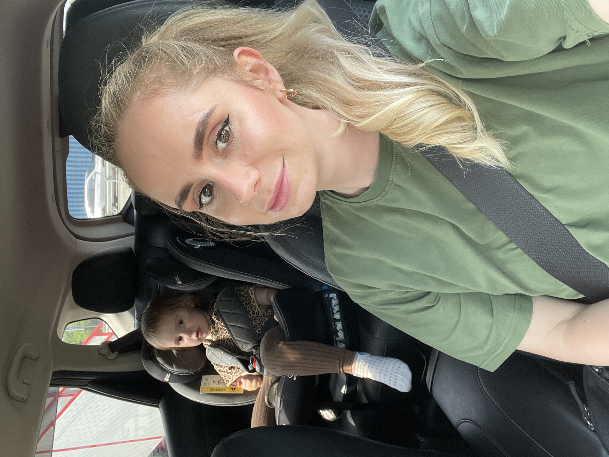 A woman takes a selfie inside a car, with a young child visible in a car seat behind her. The image is captured from an upward angle, showing the woman's face and upper torso in the foreground, turned slightly towards the camera. She has long blonde hair, styled partly in a ponytail, and is wearing a plain green t-shirt and a dark seatbelt across her chest. She has makeup on, including winged eyeliner, and a neutral expression with a slight smile. A small golden hoop earring is visible on her left ear.
Behind her, to the right, a toddler is seated in a dark-colored car seat. The child is wearing a leopard-print top, ribbed brown leggings, and white patterned socks. They are looking forward, with a neutral expression. A yellow warning label with the word "AIRBAG" is visible on the side of the car seat.
The car's interior features dark upholstery. Through the car windows, exterior elements are faintly visible, including what appears to be a structure with red and white components, suggesting a daytime setting. The scene takes place in Kiparisovo-2, Russia.
