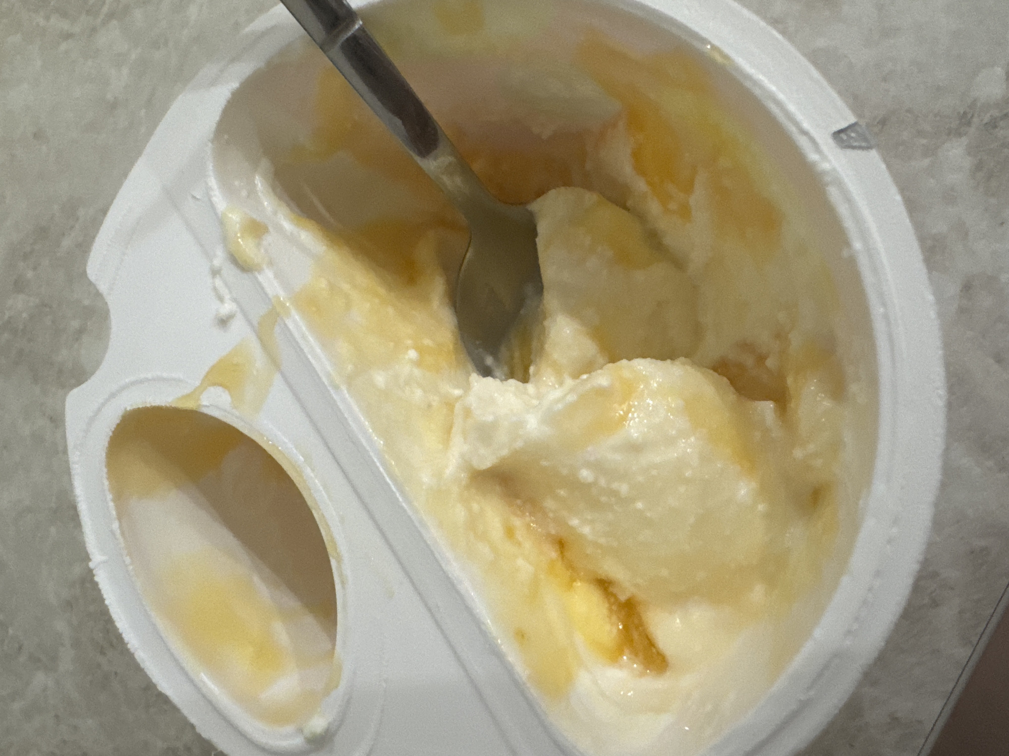 A close-up, overhead shot shows a white plastic container of yogurt with a dollop of yellow fruit or jam swirled into it. A silver spoon is partially submerged in the yogurt, as if someone is about to eat it. The container is divided into two sections, one of which appears to be empty. The yogurt is creamy and textured, with some small bubbles visible on its surface. The background is a neutral gray countertop. There is no indication of the location in Saratov, Russia.