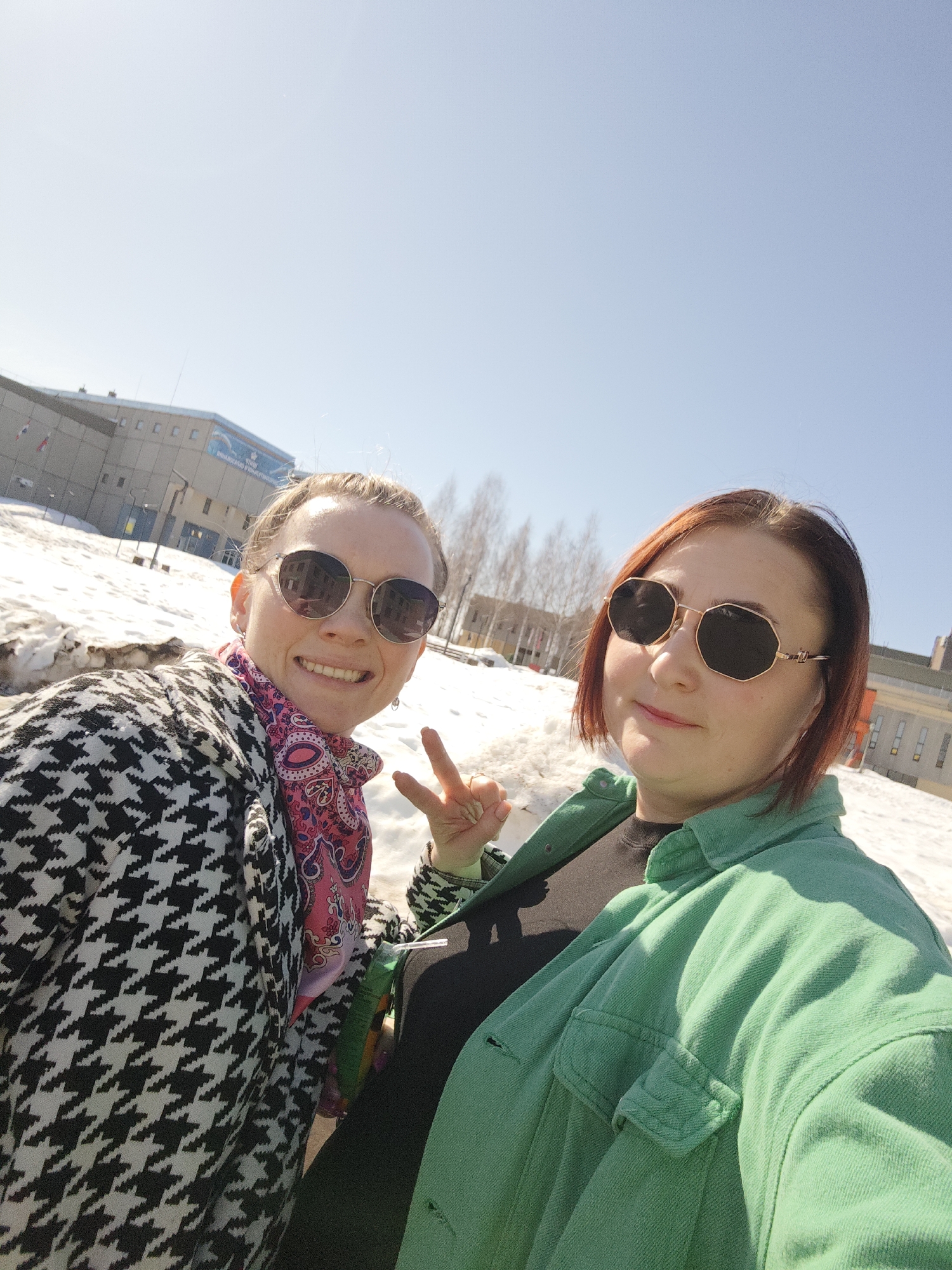 Two women wearing sunglasses are posing for a selfie outdoors. The woman on the left wears a black and white houndstooth-patterned coat and a pink patterned scarf and is making a "peace" sign. The woman on the right wears a green jacket. Snow is on the ground, and buildings are visible in the background. The location is Марковский, Russia.