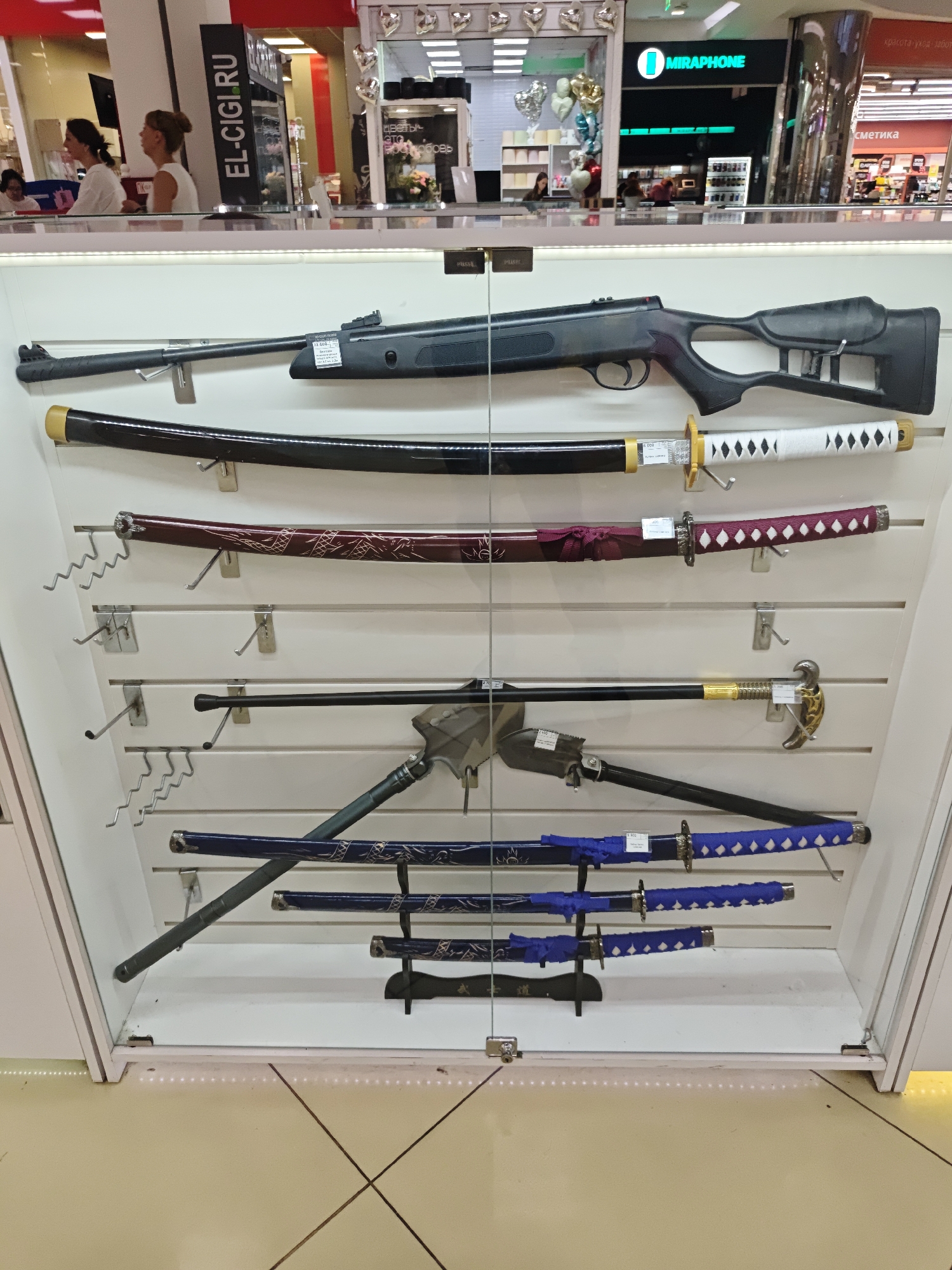 Here is a detailed description of the image:
The image captures a display case containing an assortment of weapons, including what appears to be an air rifle, several swords of various designs, and a combination shovel-axe tool, possibly for camping or survival. The items are arranged vertically within the case, mounted on a pegboard-style wall.
Background:
The display case is situated in what seems to be a retail environment, likely within a shopping mall. Visible through the glass are elements of other stores, including signage for "MIRAPHONE" and "EL-CIGIRU." People are visible in the background, some appearing to be customers or store employees. The background suggests the location is Yekaterinburg, Russia.
Subjects/Objects:
- Air rifle: A black air rifle is placed at the top.
- Swords: There are several swords of varying styles and colors, possibly decorative or replicas. The swords feature different handle and blade designs, with some having sheaths.
- Combination Tool: A folding shovel/axe tool is visible, with both the shovel and axe heads distinct.
People:
There are several individuals visible in the background, suggesting a busy commercial environment. They appear to be going about their activities within the retail setting.
Environment:
The retail setting suggests the purpose of the weapons is likely for sale or display. The area appears well-lit and organized, typical of a commercial establishment. The surroundings suggest a modern shopping mall or similar retail space.