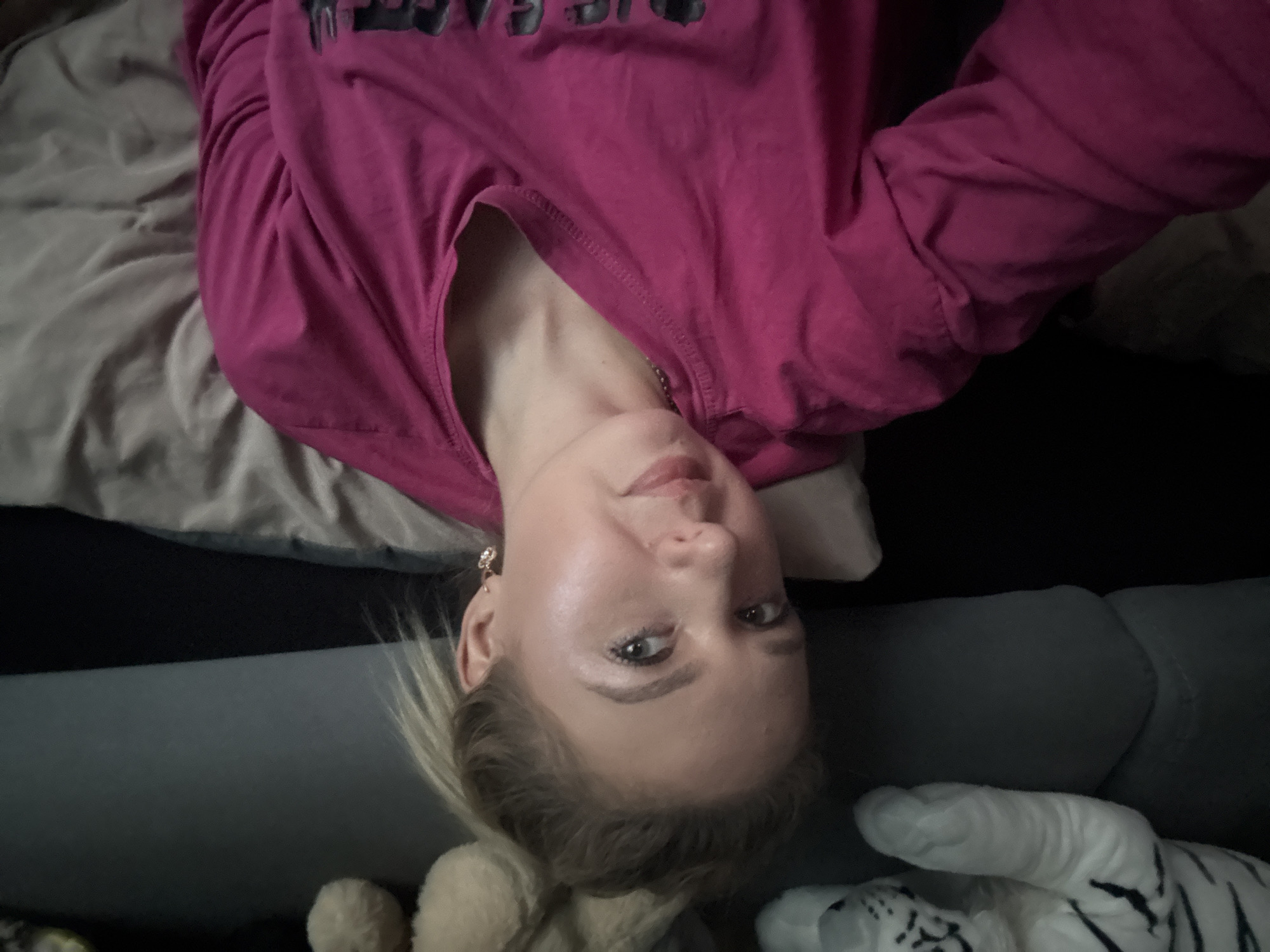 A young woman with blonde hair pulled into a messy bun is captured in a selfie, lying down. She is wearing a bright pink long-sleeved shirt. Her head is tilted slightly, and she is looking towards the camera with a relaxed expression. A faint smile plays on her lips, and she is wearing a small, dangling earring in her visible ear. The background consists of a dark grey couch and a muted, wrinkled fabric, possibly a blanket or bedsheet. To the left of the woman, a portion of a stuffed white tiger with black stripes is visible, its paw resting on the couch. To the right of her head, the fuzzy ears of a stuffed animal are apparent. The lighting appears dim, creating a soft, intimate atmosphere.