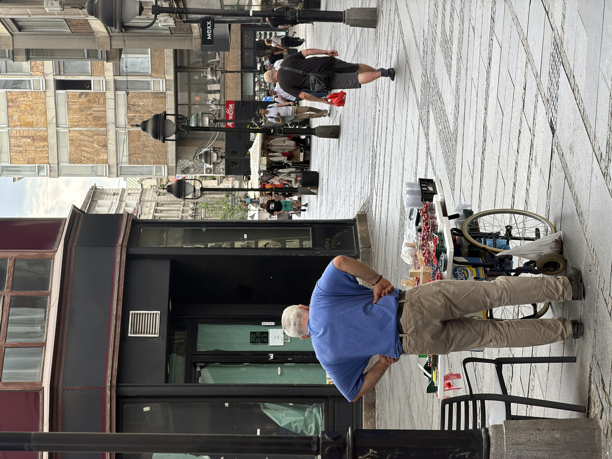 The image captures a street scene in Belgrade, Serbia, likely on a pedestrianized shopping street given the architecture and the presence of street vendors and shops. In the foreground, a man with graying hair, wearing a blue t-shirt and tan pants, stands with his hands on his hips, facing a mobile cart laden with berries. The cart has a bicycle wheel attached to one side. Behind the man, a dark-colored building with large windows and what appears to be a closed shopfront dominates the left side of the frame. The pavement is made of stone tiles laid in a patterned design. Further down the street, other people are visible walking, and shops with awnings and signs, including "MEXX," line the street. The buildings are varied in architectural style, suggesting an urban environment with a mix of older and newer constructions. The lighting indicates it is daytime, likely overcast, as there are no strong shadows.