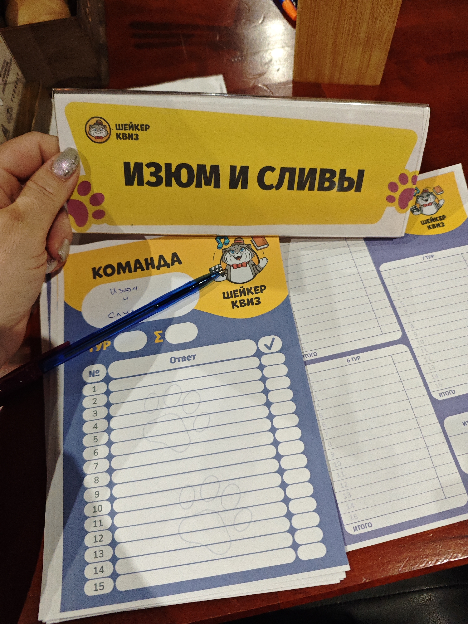 The image shows a table setting for a quiz competition. On a dark wooden surface, a person's hand is holding a sheet labeled "Шейкер Квиз" (Shaker Quiz) with the team name "Изюм и сливы" (Raisins and Plums). Below this sheet is the answer sheet, with numbers 1-15 for the answers. A blue pen is placed on the sheet. Another sheet with columns for multiple rounds of the quiz (6 ТУР, 7 ТУР) is visible to the right. The presence of "Шейкер Квиз" signage indicates the location of the competition could be in the city of Chaikovsky, Russia.