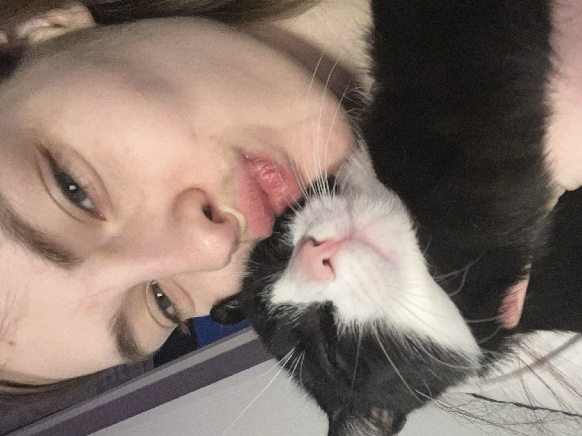 A close-up, selfie-style image depicts a person with fair skin and light brown hair holding a black and white cat. The person's face is partially visible, showing one eye with eyeliner, a nose, and lips. The cat's face is prominent, featuring a pink nose, white fur around the mouth, and black fur on the head and body. The background is indistinct, but a dark blue and gray surface is visible. The orientation of the image is rotated, with the person and cat appearing sideways. No elements suggest a location in Saint Petersburg, Russia.