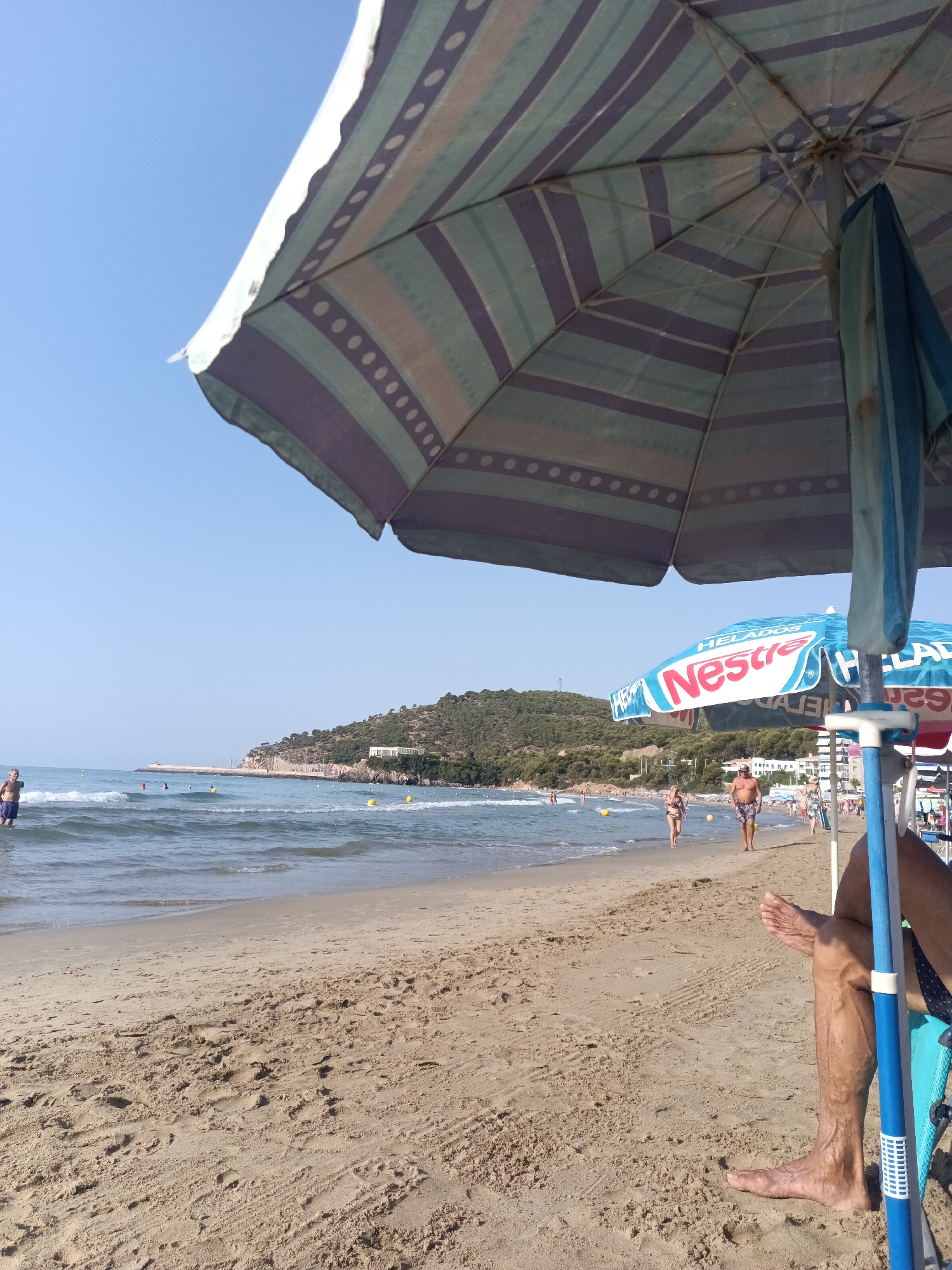A person's legs and feet are visible in the foreground, resting on sandy ground. The person is sitting under a large, striped umbrella. In the middle ground, a beach stretches along the coastline. Several people are walking along the shore, and others are in the water. A second umbrella, advertising "Nestle Helados", is also visible. The background features a hill covered with trees, and buildings can be seen on the hillside. The sea is calm with small waves. The sky is clear and blue. The location is likely a beach in La Playa, Spain, given the context provided.