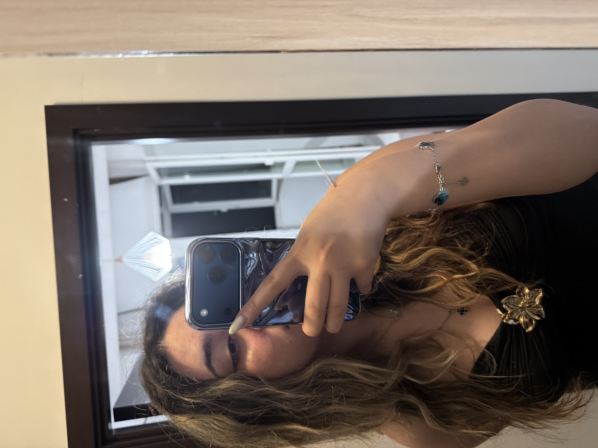 A young woman with long, wavy blonde hair is taking a mirror selfie in what appears to be a home interior. She is wearing a black, ruched top and a delicate silver bracelet with blue and heart-shaped charms on her left wrist. Around her neck, she has a silver chain with a small, dark cross pendant and a larger, ornate gold flower-shaped pendant on her chest. Her right hand holds a dark, reflective phone, angled to capture her reflection. The background, visible in the mirror, shows a room with a large window with white blinds or shutters, and a modern-looking pendant light fixture hanging from the ceiling. The room has pale walls and wooden trim around a doorway. The overall setting suggests a private residence. There is no visual information in the image to suggest a specific location in Plovdiv, Bulgaria.