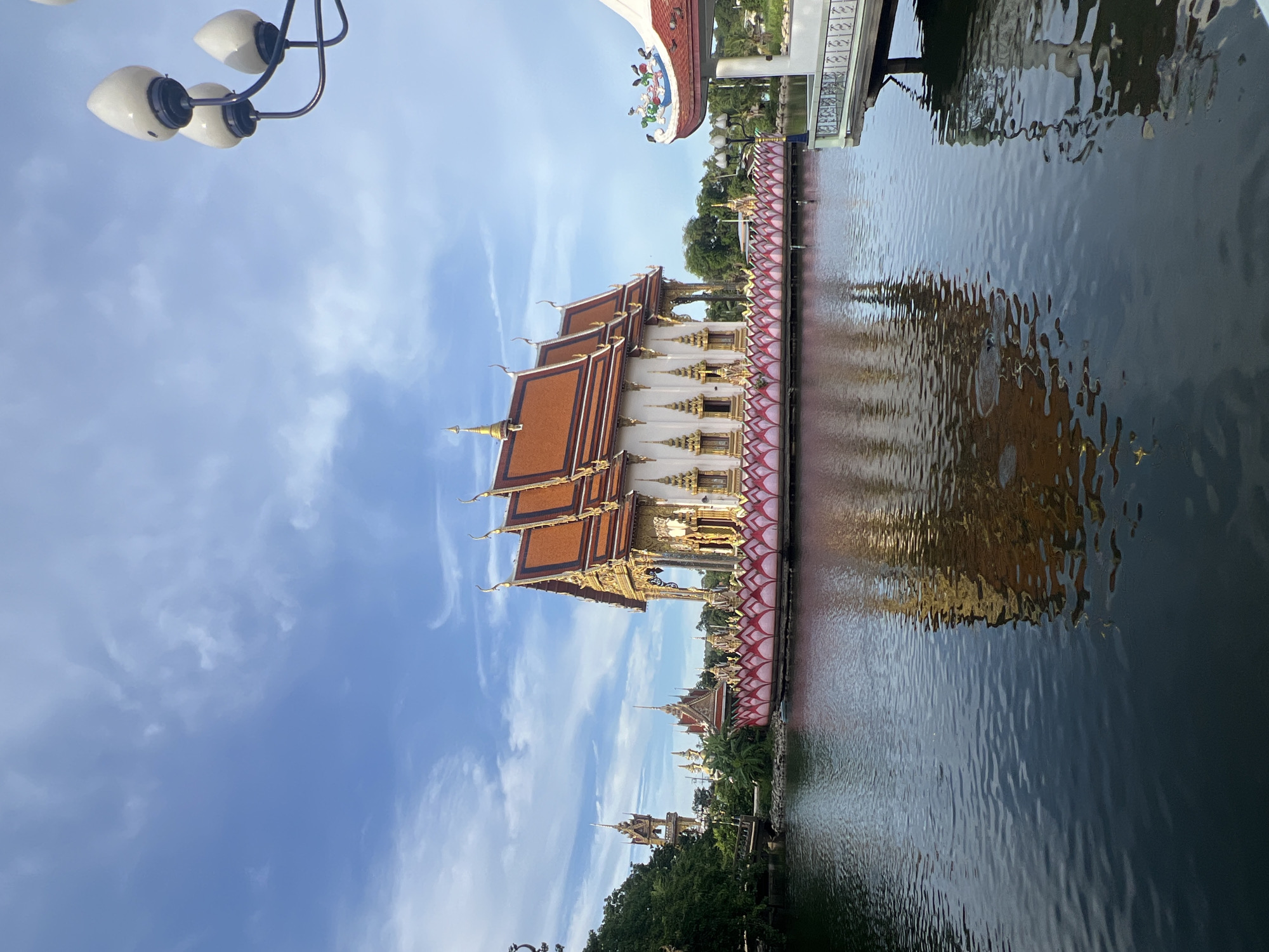 The image shows a temple complex in Baan Plai Laem, Thailand. The main structure is a white building with a golden, multi-tiered roof, situated on a body of water. The building is surrounded by lotus-shaped decorations in pink and gold. Several other temple spires and structures are visible in the background, along with lush green trees. The sky is blue with scattered white clouds. The water reflects the temple and the sky. In the foreground on the right, a decorative lamp post with three white, pointed lights is visible against the sky. Also on the right is a white balcony with a decorative railing and a red-tiled roof adorned with colorful figures. The overall scene depicts a serene and ornate religious site.