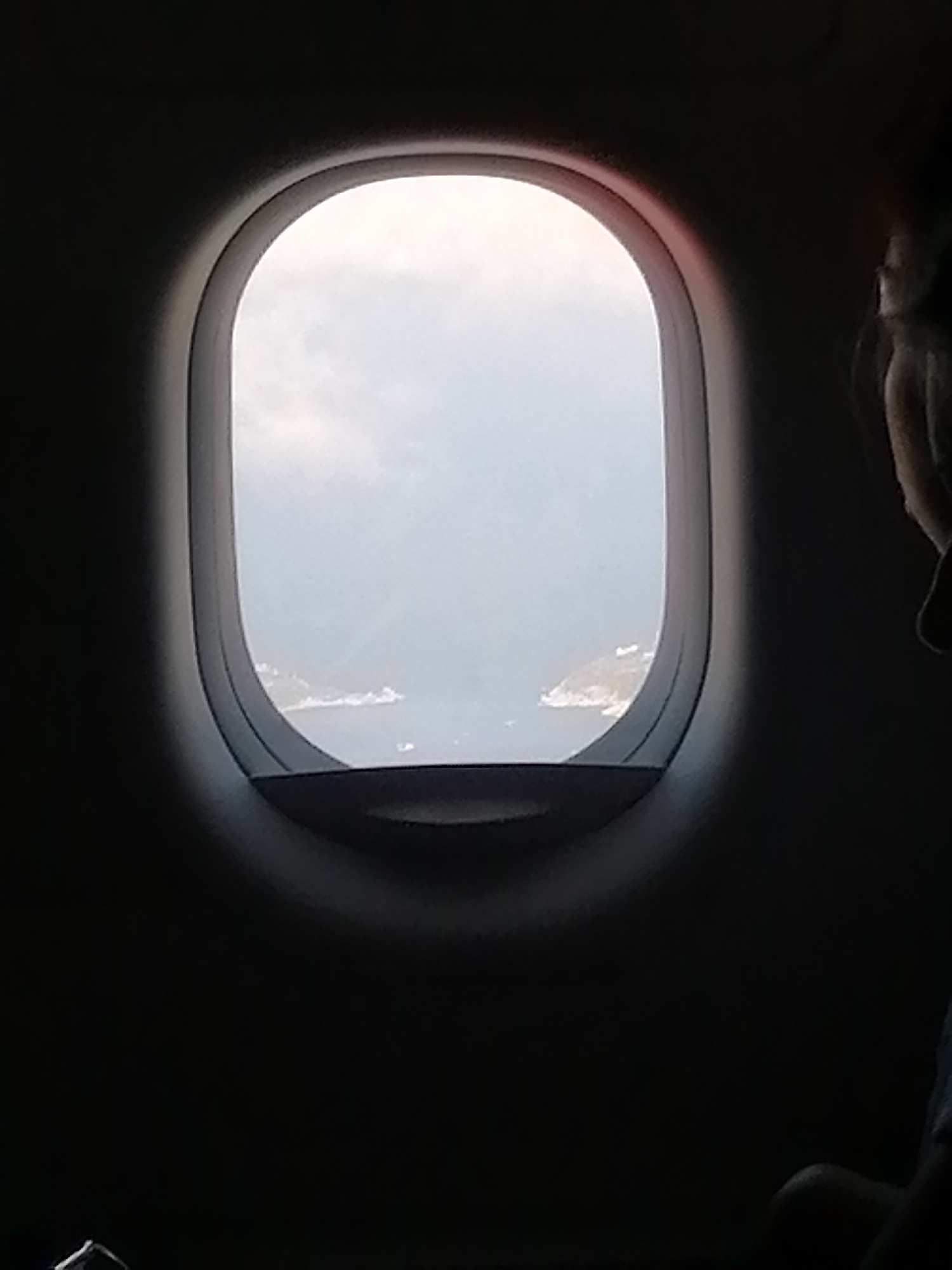 The image shows a view from an airplane window. Outside the window, there is a body of water with landmasses on either side. The sky above the water is light. The airplane window frame is visible, and part of a person's head and shoulder are visible on the right side of the image. The location is Unknown, Unknown.
