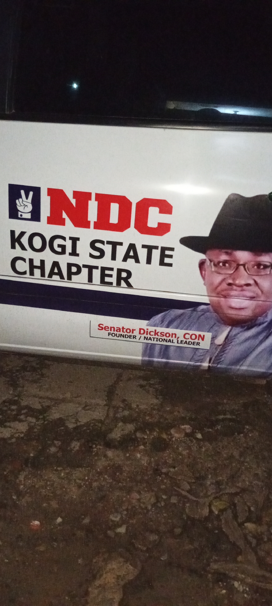 A photo of a vehicle with the political banner of the NDC (National Democratic Congress) Kogi State Chapter showing the image of Senator Dickson, labeled as founder/national leader, is depicted against a dark background. The photo appears to be taken in Adankolo, Nigeria, judging by the Kogi State Chapter affiliation on the banner.