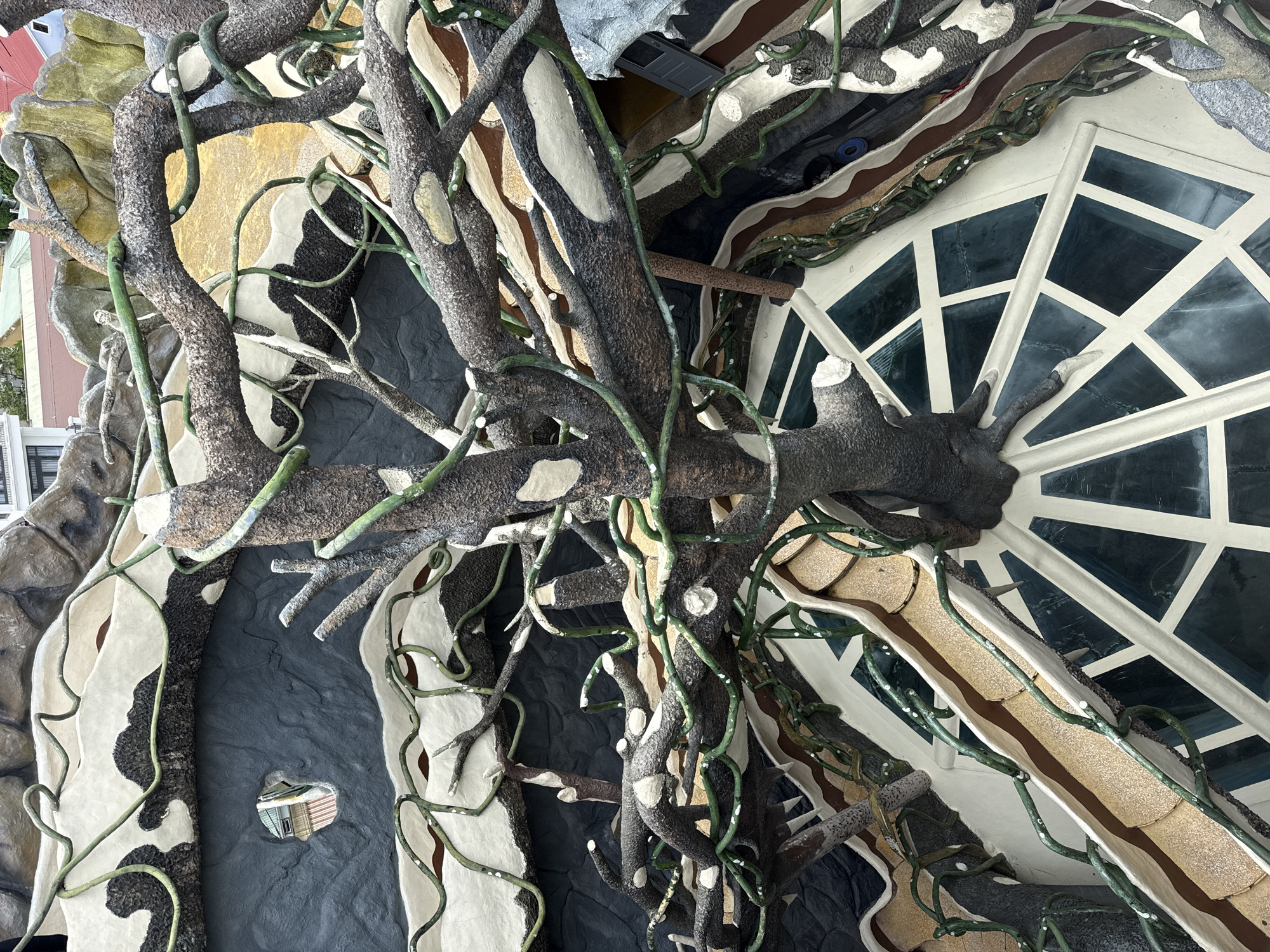 The image captures an unconventional architectural detail of Hang Nga guesthouse, also known as the Crazy House, in Da Lat, Vietnam. A prominent feature is a structure resembling a gnarled tree, with thick, dark branches and green vines adorned with white spots twisting and turning across the scene. The tree-like structure is integrated into the building's facade. The building has a white frame with a glass-filled, diamond-patterned grid. The surfaces surrounding the tree are irregular, with alternating sections of dark gray and textured white, creating a cave-like opening. A tiny hole is visible in the dark gray section, offering a distorted reflection of the exterior. Above the tree is a section decorated with horizontal bands of brown, white, and dark gray, with some mechanical-looking gray objects visible near the top of the frame.