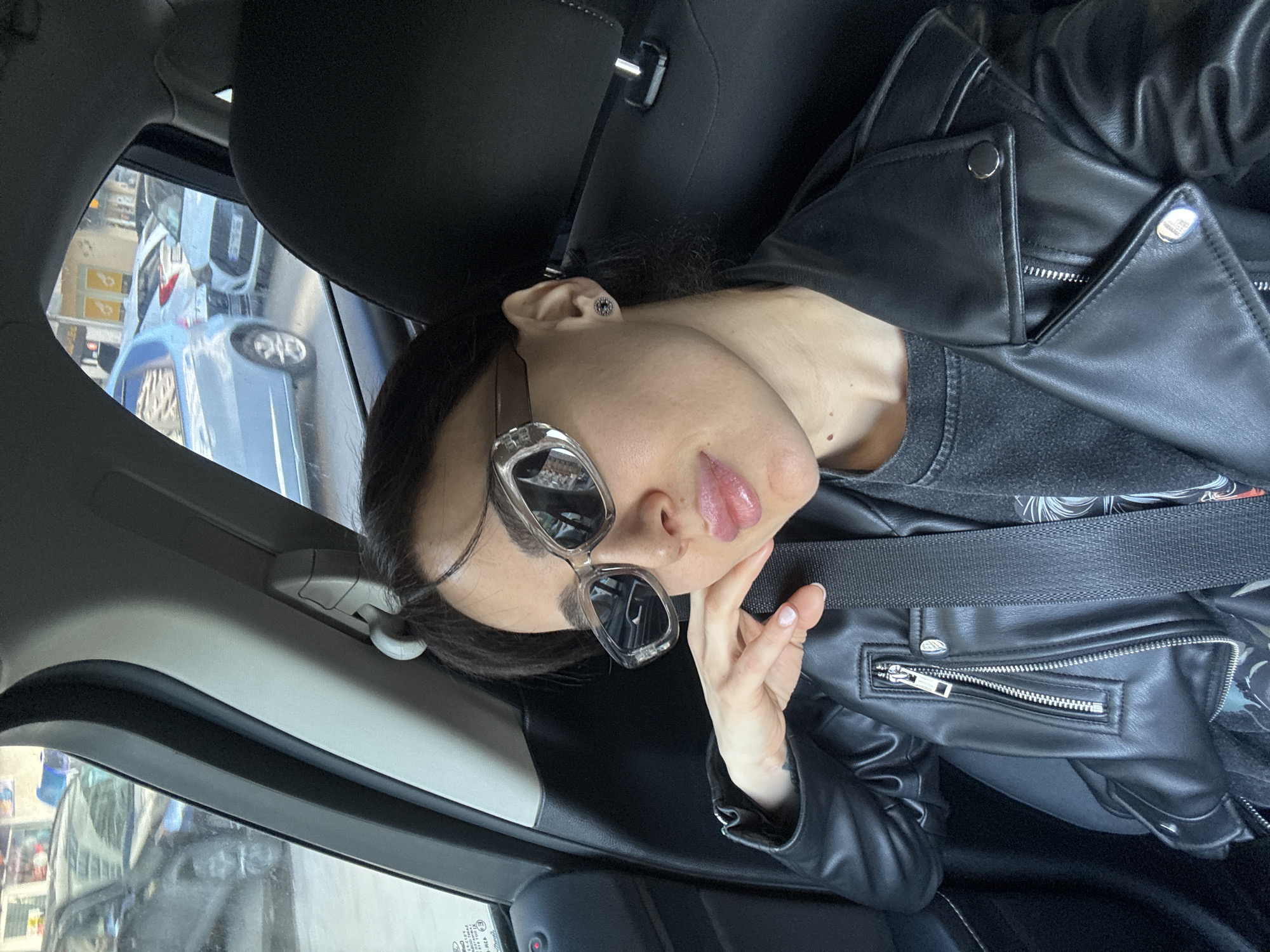 The image shows a woman wearing a black leather jacket and sunglasses in a car, likely a passenger seat. She has on a black shirt, and a seatbelt is visible across her chest. The background visible through the window shows a city scene, possibly in Tbilisi, Georgia, with cars and a glimpse of buildings.
