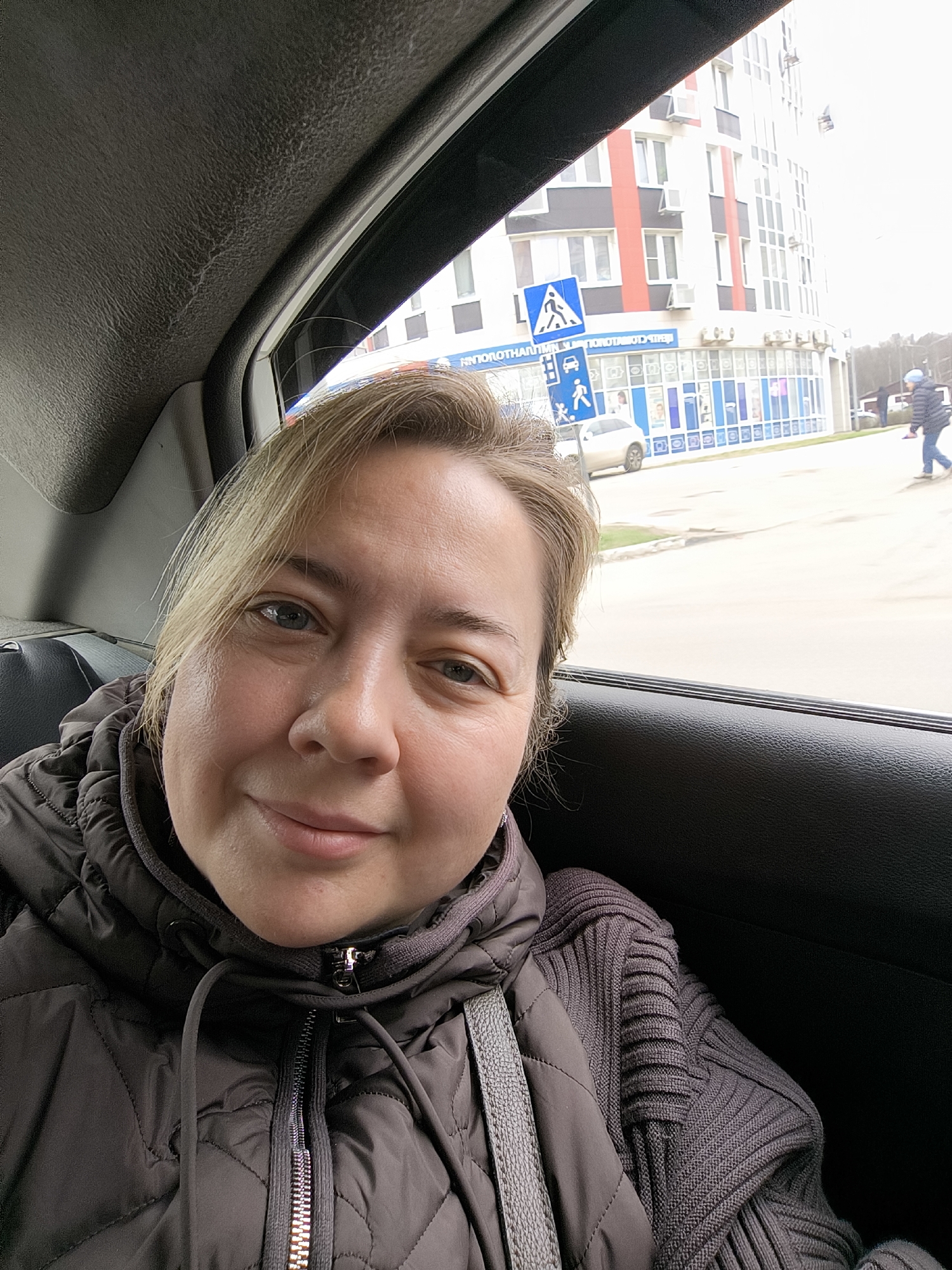 The image depicts a woman in the back seat of a car, presumably traveling through the city of Khimki, Russia. The woman is wearing a brown quilted jacket over a knitted sweater. Outside the car window is a multi-story building with white, grey, and red accents, and a store underneath with a sign in Russian. A crosswalk sign is also visible. A pedestrian is walking on the sidewalk in the background. The sky appears overcast.