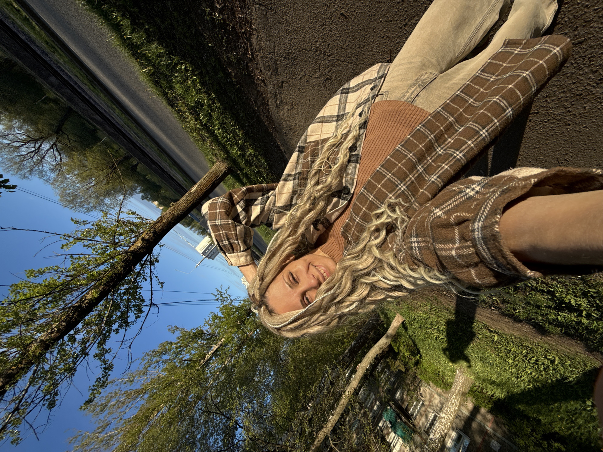 A young woman with long blonde dreadlocks is reclining on a patch of dark soil and asphalt, potentially a roadside shoulder or the edge of a path. She is smiling and appears to be taking a selfie. She is wearing light-colored jeans, a brown turtleneck sweater, and a brown and beige plaid flannel shirt. In the background, there is a tree with branches reaching towards a blue sky. Beyond the tree is a street and what seems to be a building with green and white siding. The scene is illuminated by sunlight. While precise location information is unavailable, it is stated to be in Rusanovo, Russia.
