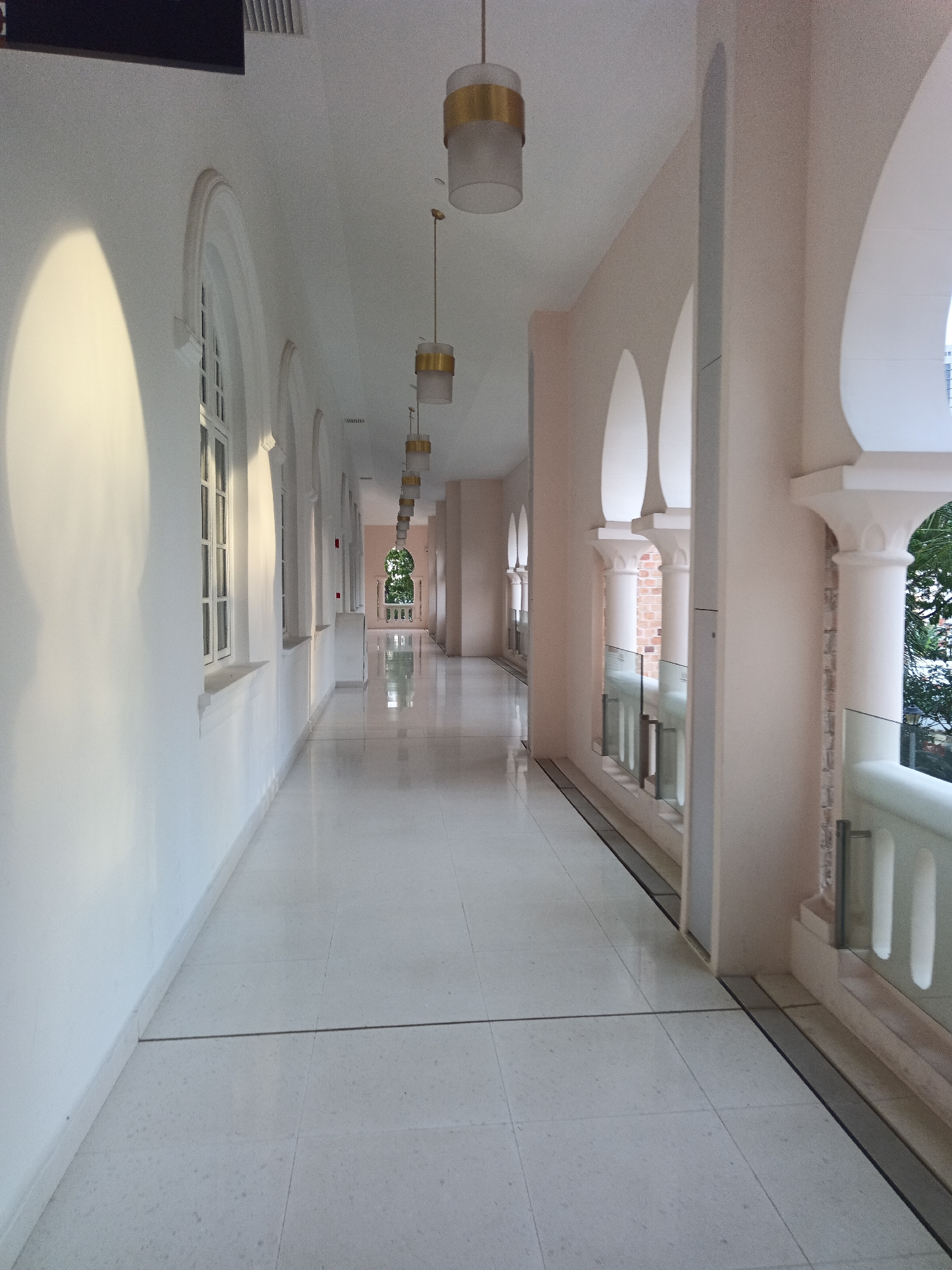 The image shows a long, brightly lit interior hallway with a white, polished floor and white walls. The hallway is lined with arched openings and columns on the right side, with glass railings visible. Several cylindrical pendant lights with gold accents hang from the ceiling. Sunlight casts a large, soft shadow on the left wall. In the distance, at the end of the hallway, an arched opening reveals greenery and an ornate railing. This hallway appears to be part of a building with architectural influences, possibly a hotel, museum, or government building. Based on the architectural style and the typical design of such structures in Kuala Lumpur, Malaysia, this hallway is likely located within the grounds of the Sultan Abdul Samad Building. The scene is devoid of any people.