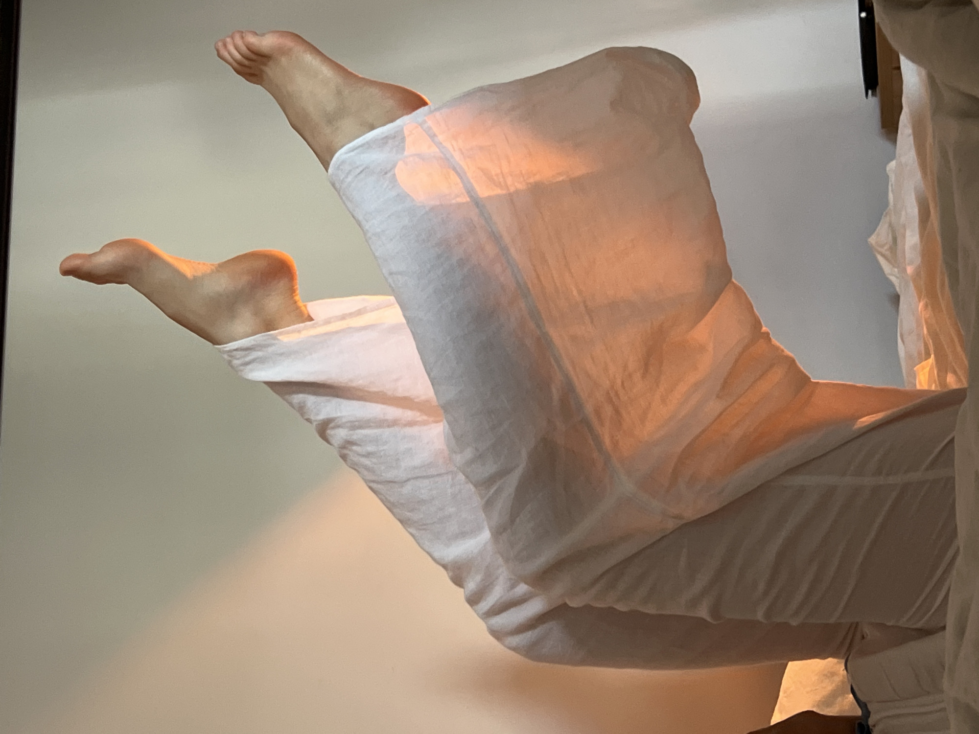 The image depicts a pair of feet, likely human, extending upwards while encased in what appears to be white linen trousers. The feet are positioned against a plain, light-colored wall. The scene is bathed in a warm, orange light. The backdrop is nondescript. There is no evidence of the location being in Ternopil, Ukraine.